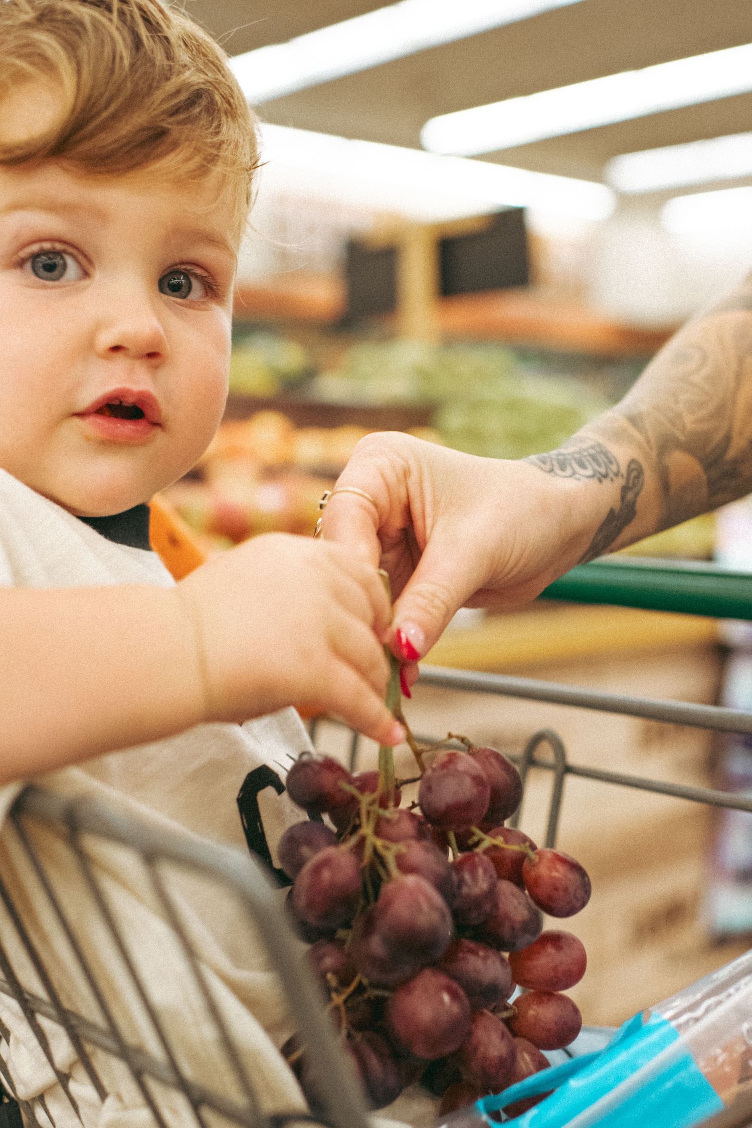 Toddler in shopping cart reaches for red grapes handed by tattooed adult in grocery store produce section.