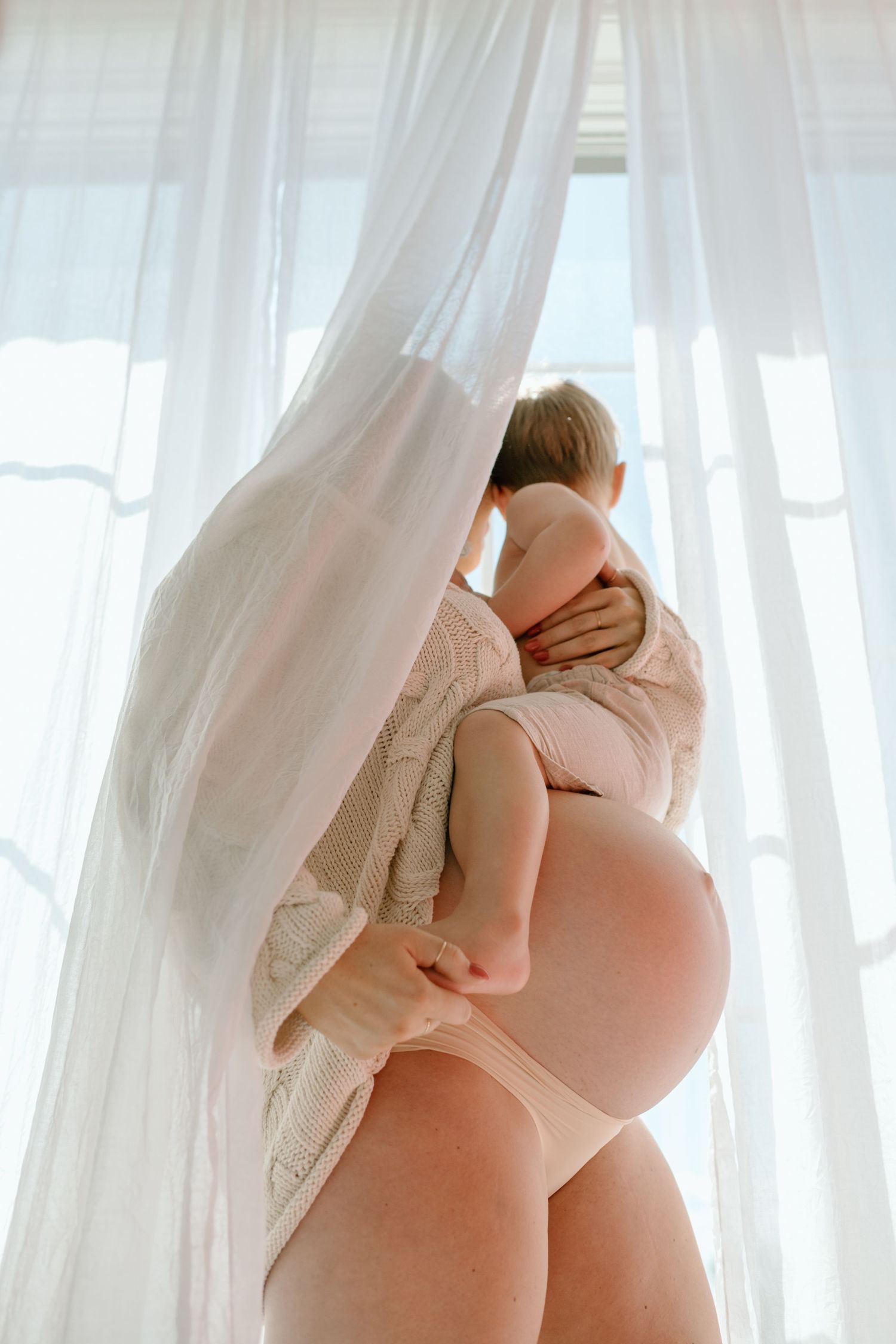 Pregnant woman in pink underwear holding a baby near sheer white curtains by a bright window.