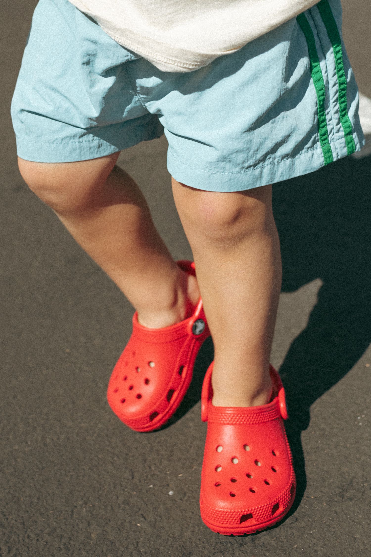 Toddler wearing red Crocs clogs and light blue shorts standing on pavement.