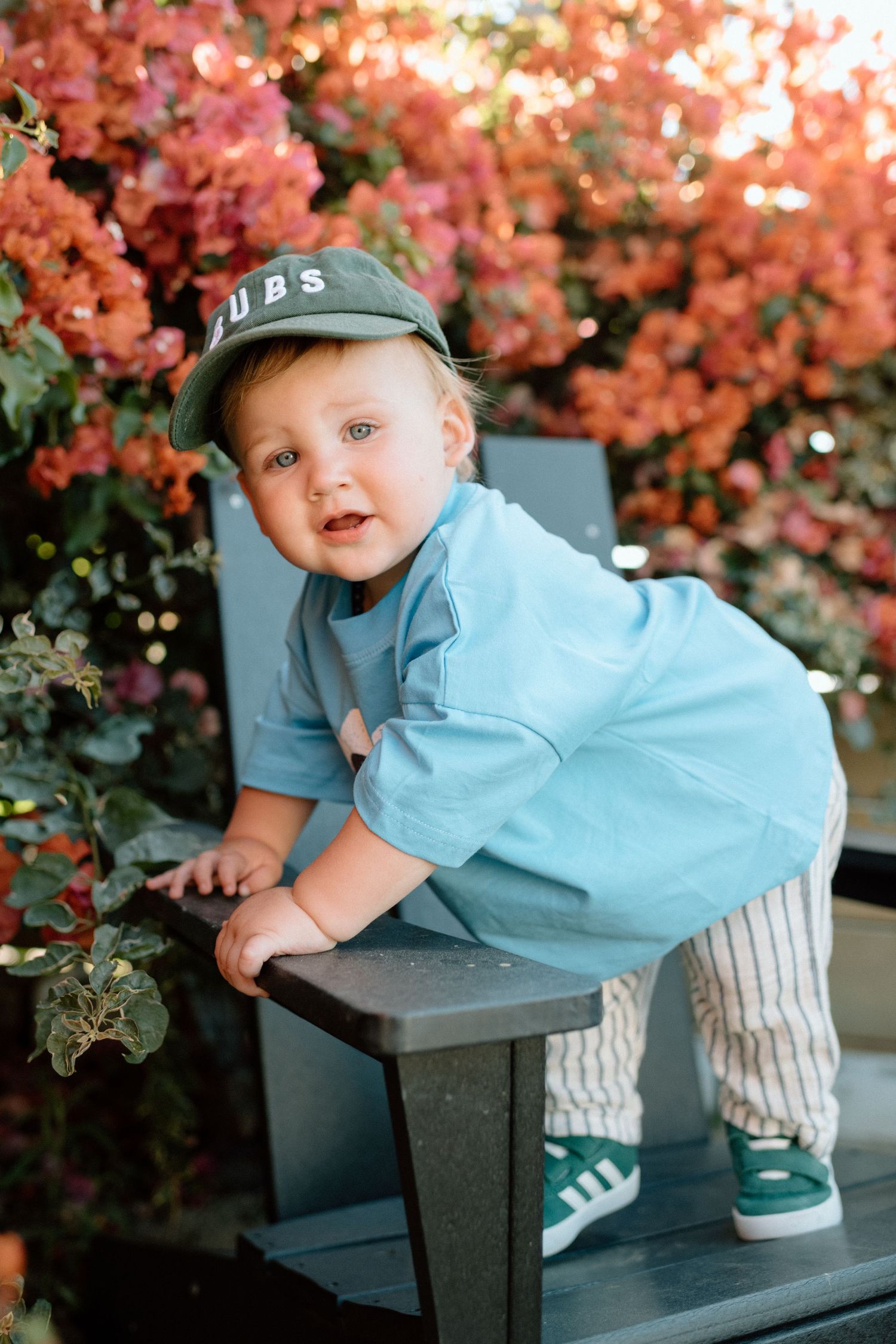 A toddler in blue clothing and baseball cap plays on outdoor furniture near blooming pink flowers.