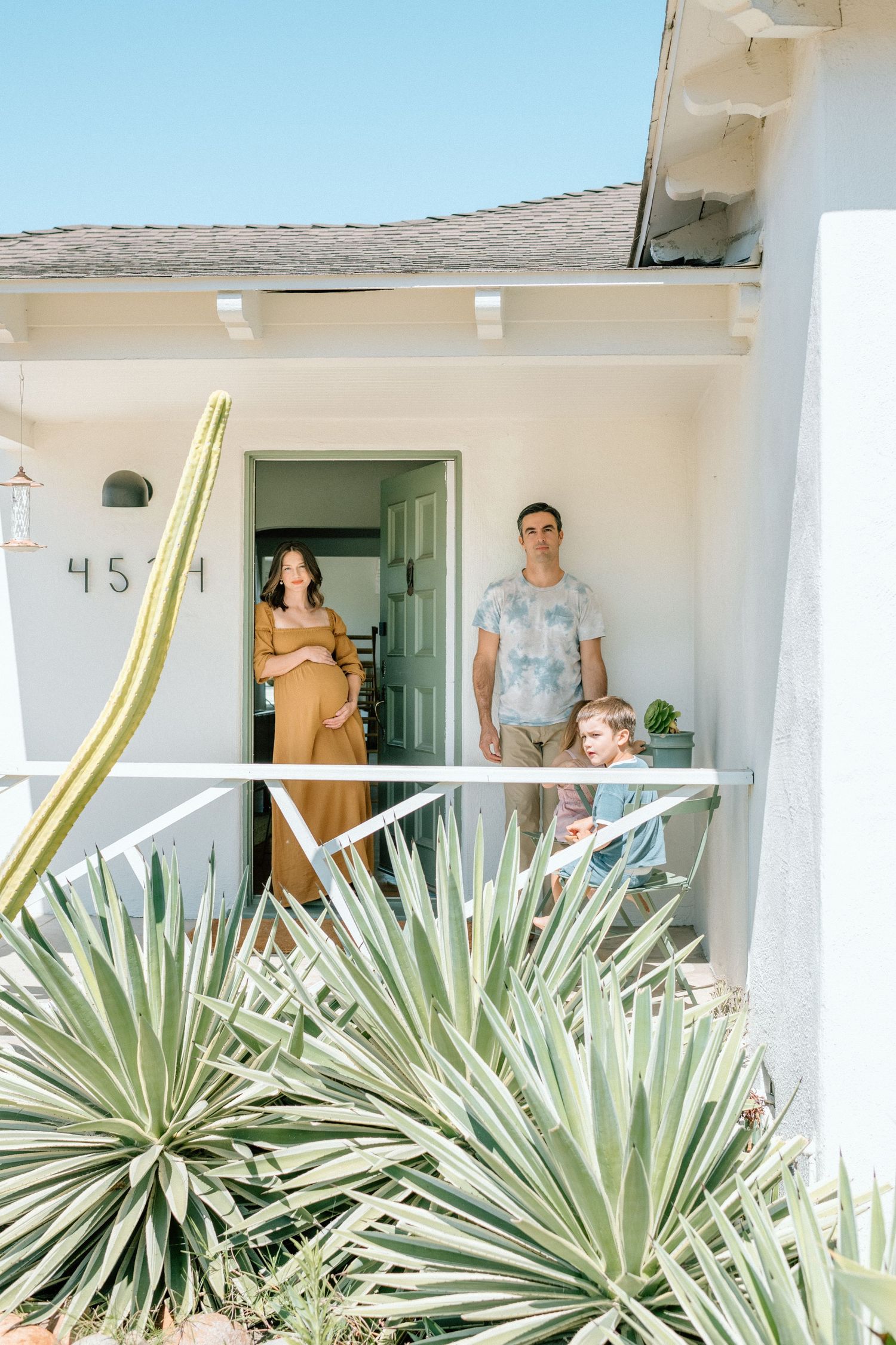 Family of four standing on a white stucco home porch surrounded by desert plants under a clear blue sky.