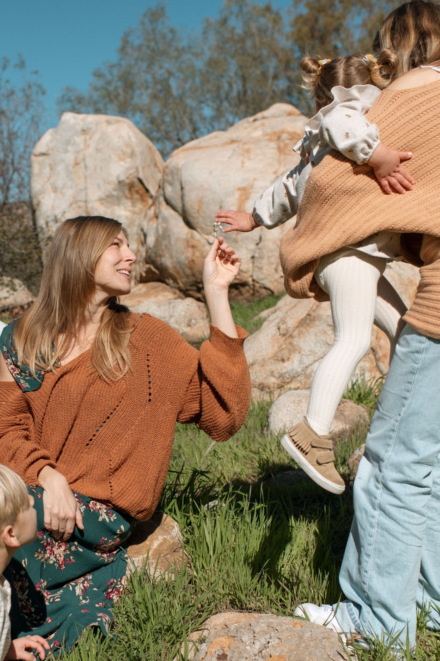A smiling mother in an orange knit sweater sits on grass while father lifts their toddler near rocks.