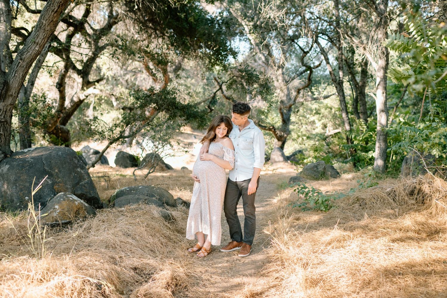 Couple admiring pregnant belly together on a sunny wooded trail with large boulders.