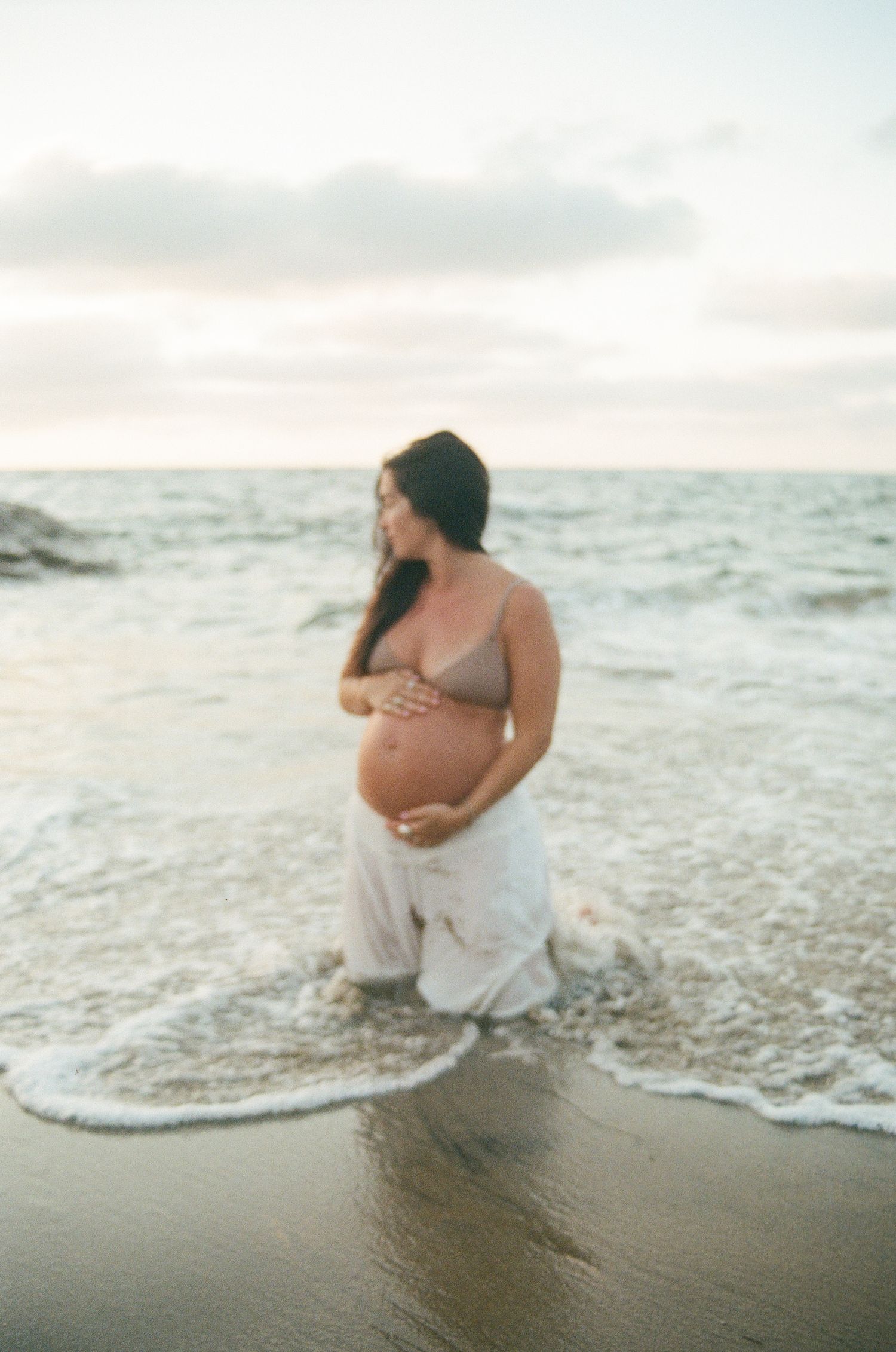 A pregnant woman in a bikini top and white pants cradles her belly while standing in ocean waves at sunset.