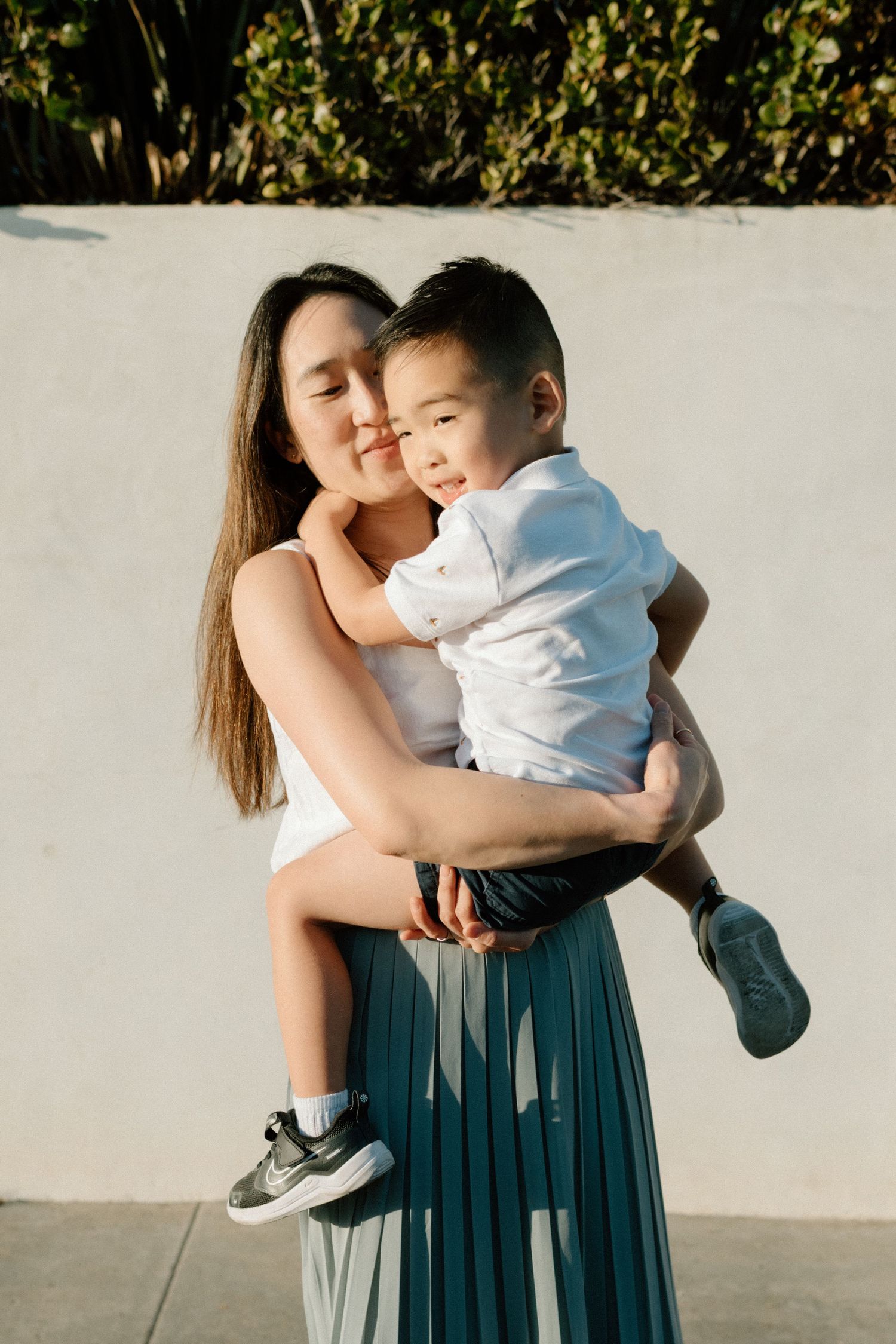 Asian mother lovingly holds young son in her arms outdoors against a white wall in sunlight.