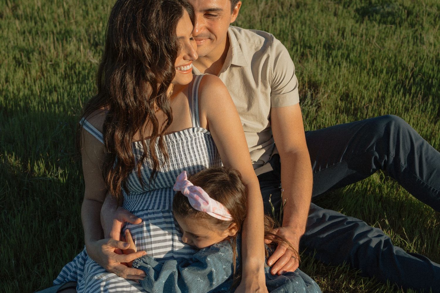 Couple sits in golden grass at sunset, pregnant woman cradled by partner as toddler leans in.