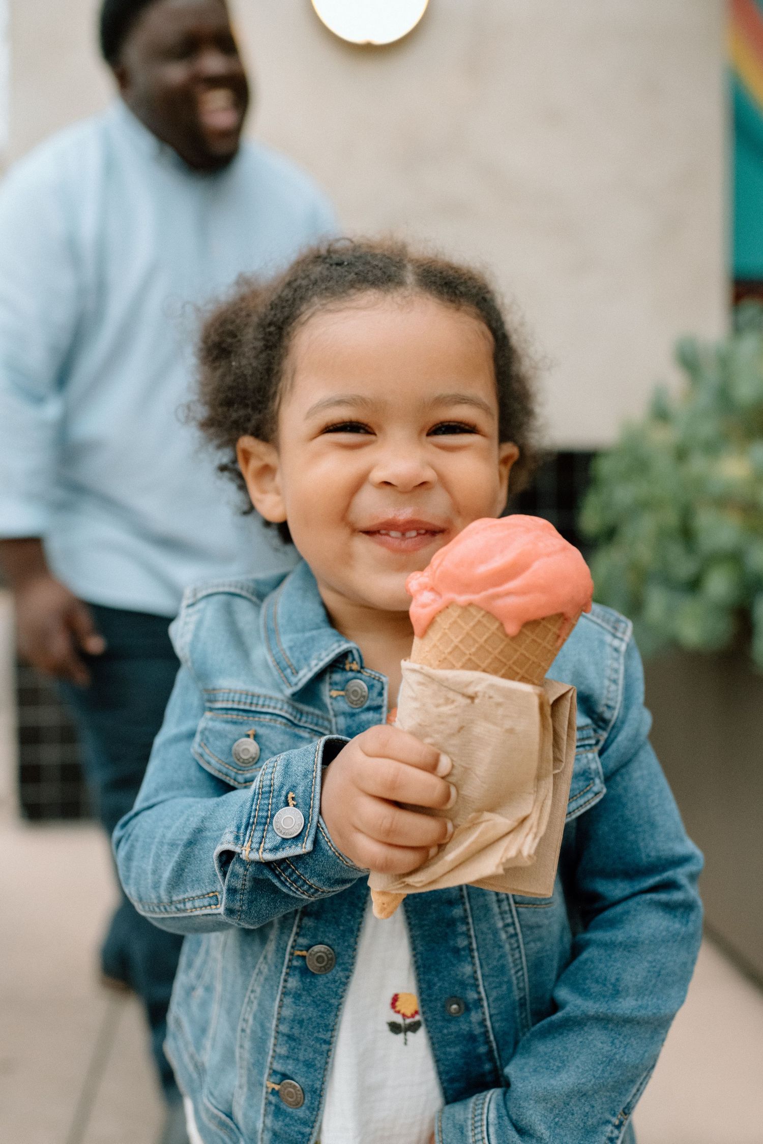 Happy young child smiling while holding a pink ice cream cone in a denim jacket outdoors.