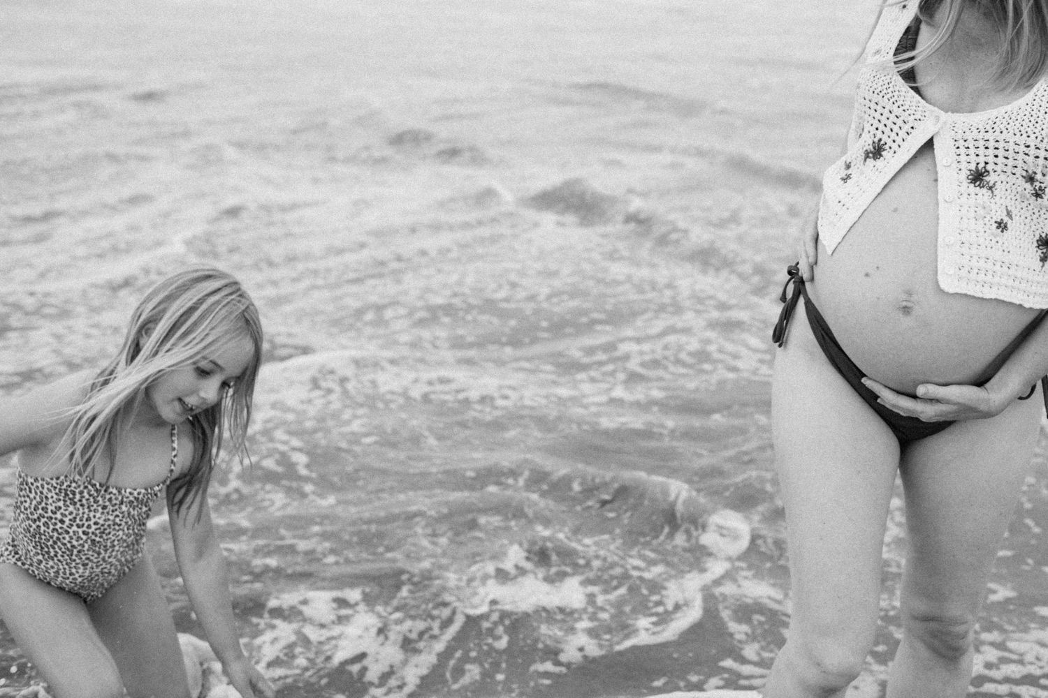Black and white photo of young girl and pregnant woman playing together at ocean shoreline.