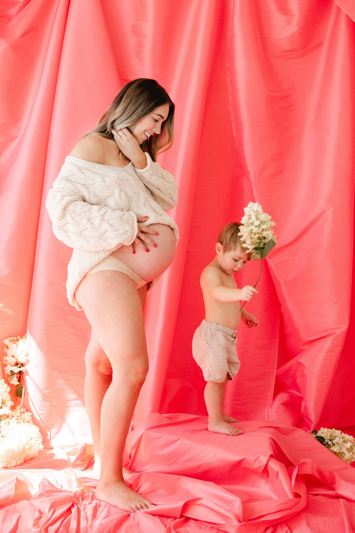 Pregnant woman in cream knit sweater smiles at toddler holding flowers against vibrant pink fabric backdrop.
