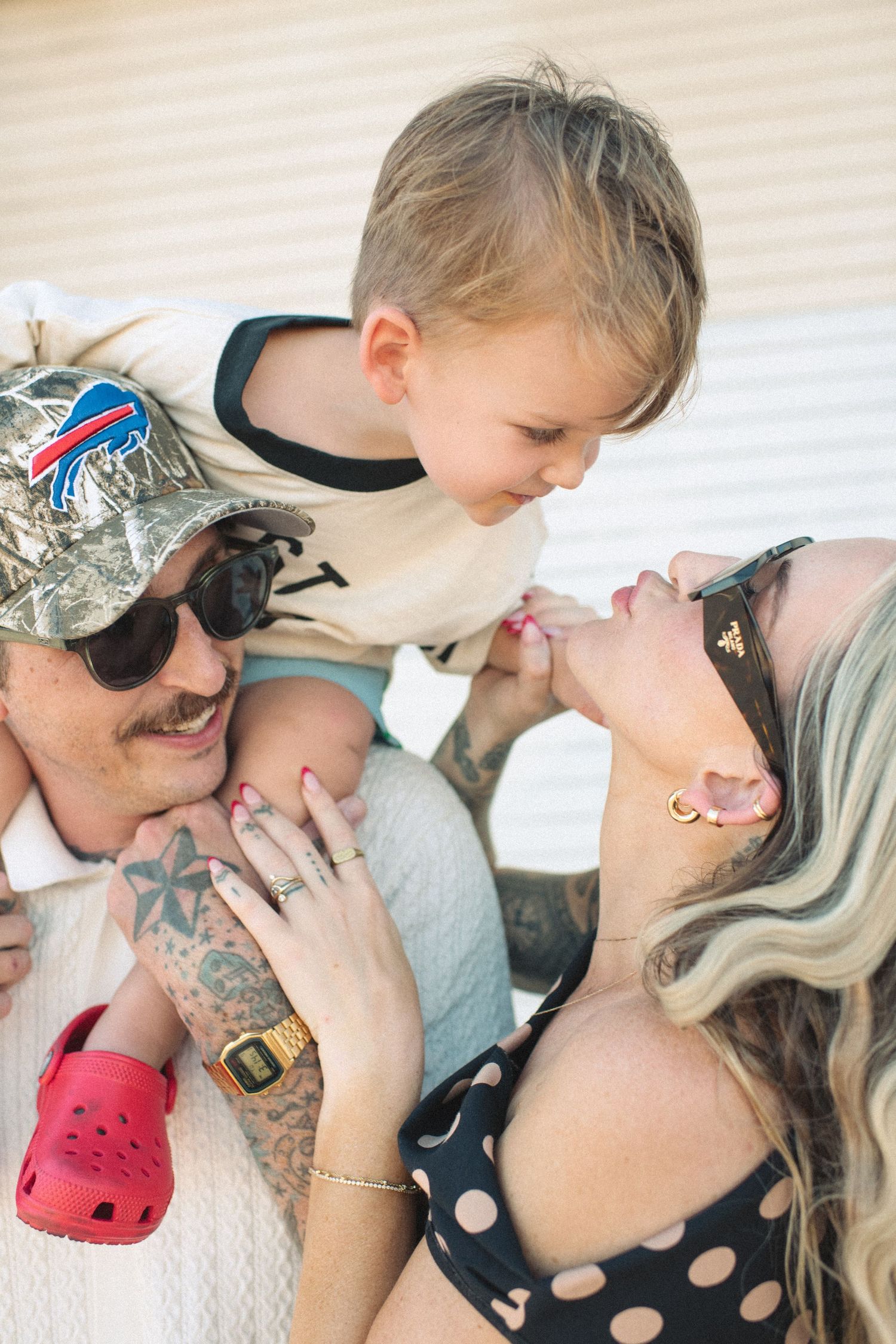 Tattooed father in Buffalo Bills cap holds laughing toddler while mother smiles nearby.
