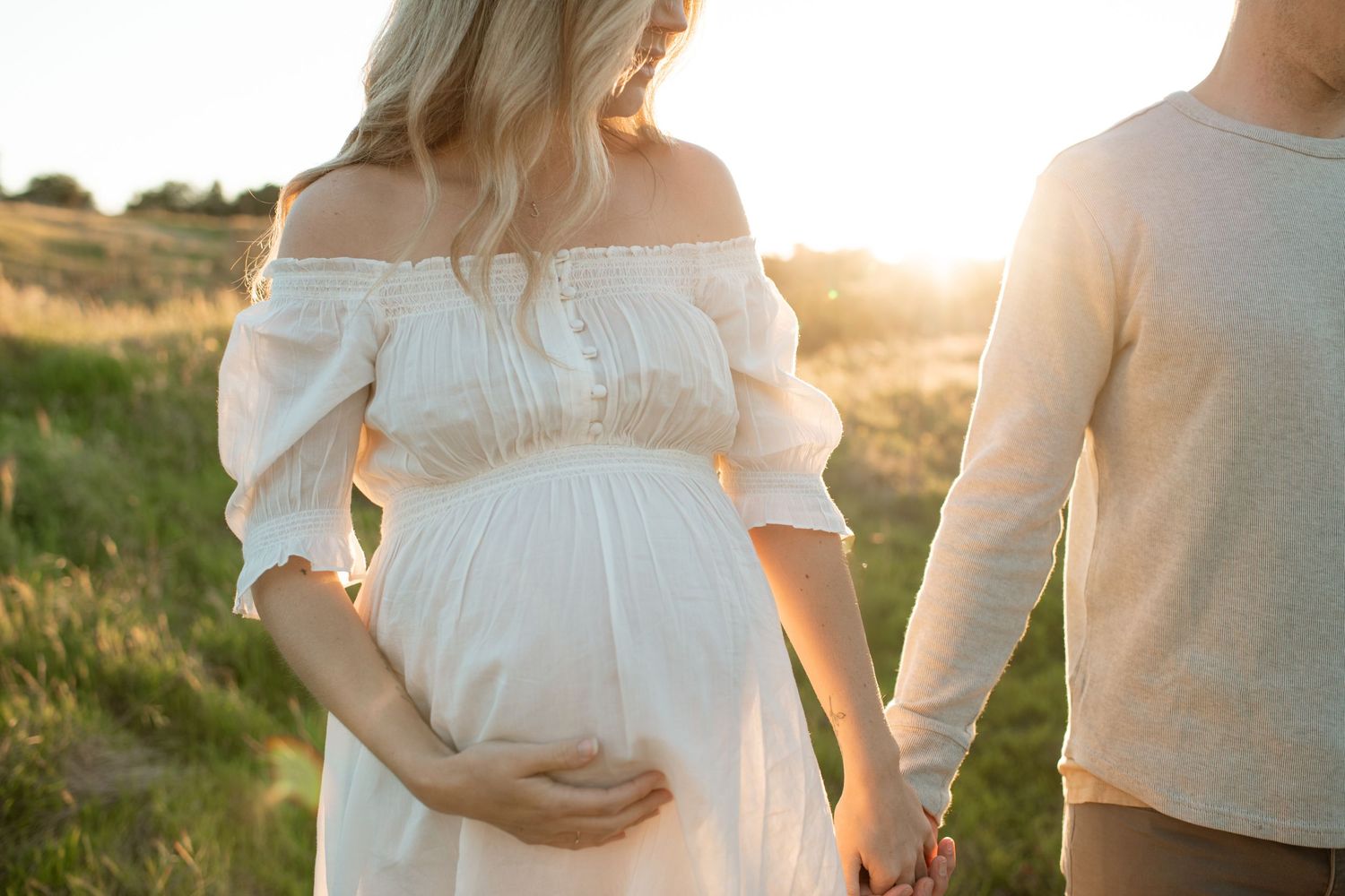 Pregnant woman in white off-shoulder dress holding belly while partner holds her hand at sunset.