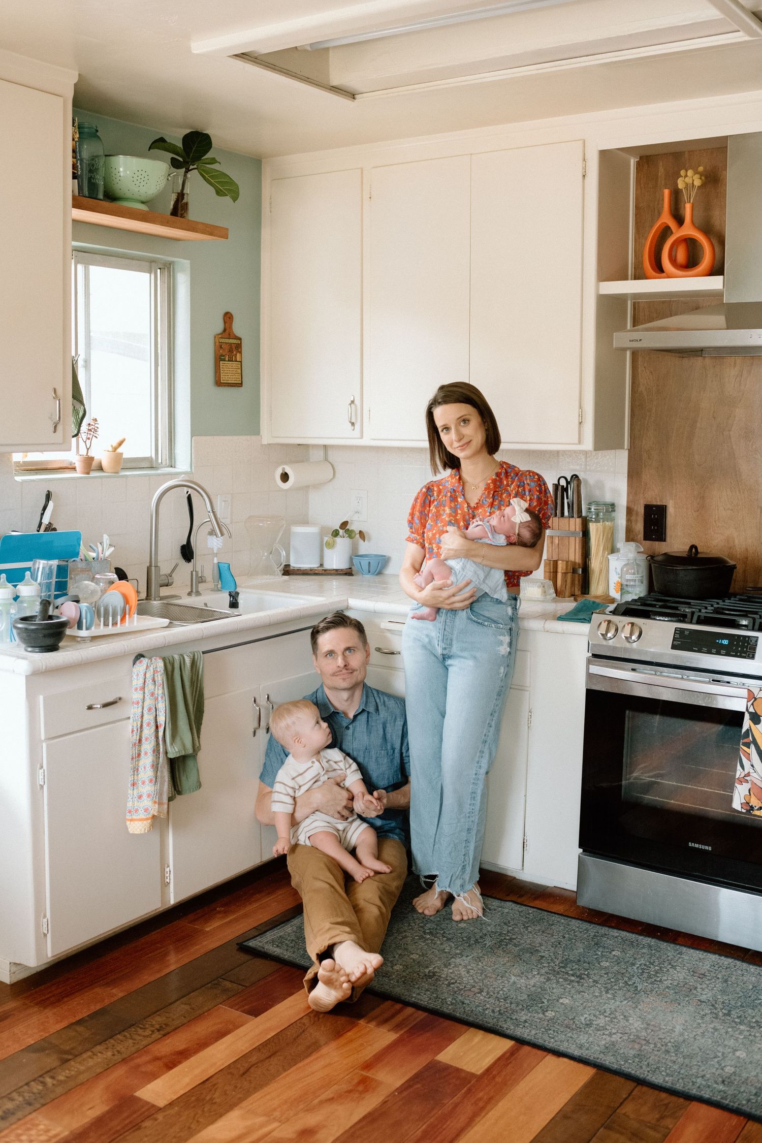 A couple with two babies poses in a bright white kitchen with hardwood floors and modern appliances.