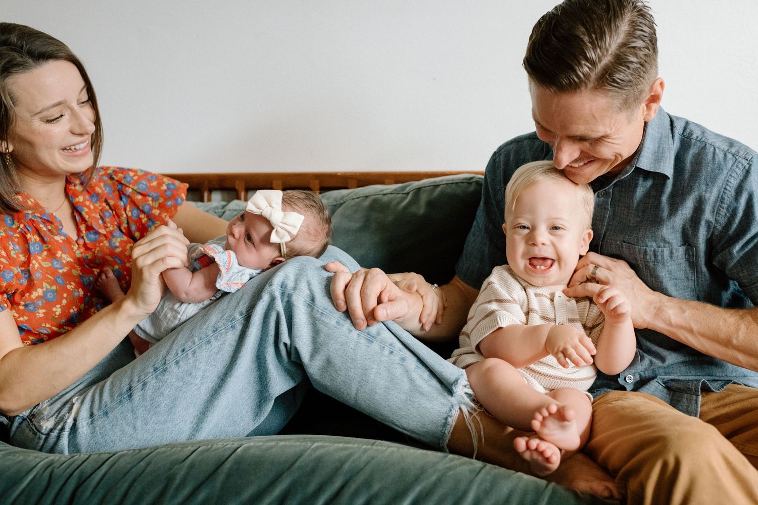 Happy family of four sits on green sofa, parents smiling at laughing toddler while mother holds newborn baby.