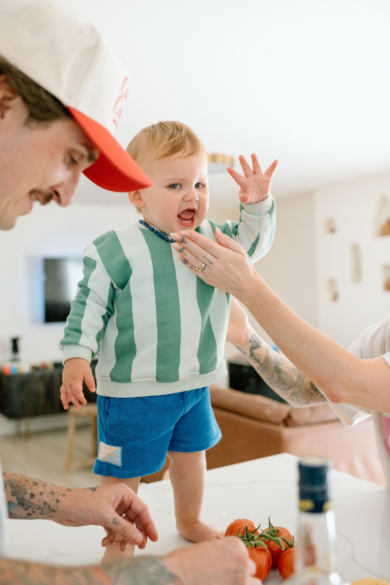 A toddler in a green striped shirt and blue shorts reacts excitedly while adults prepare food in a kitchen.