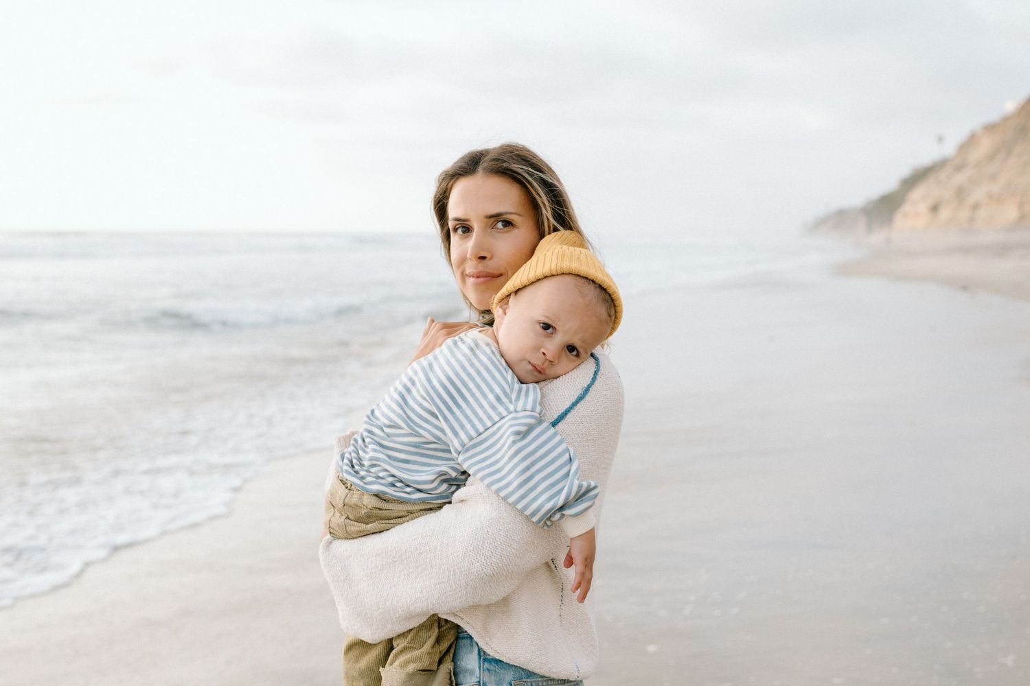 A mother holds her baby in a striped outfit while standing on a scenic ocean beach.