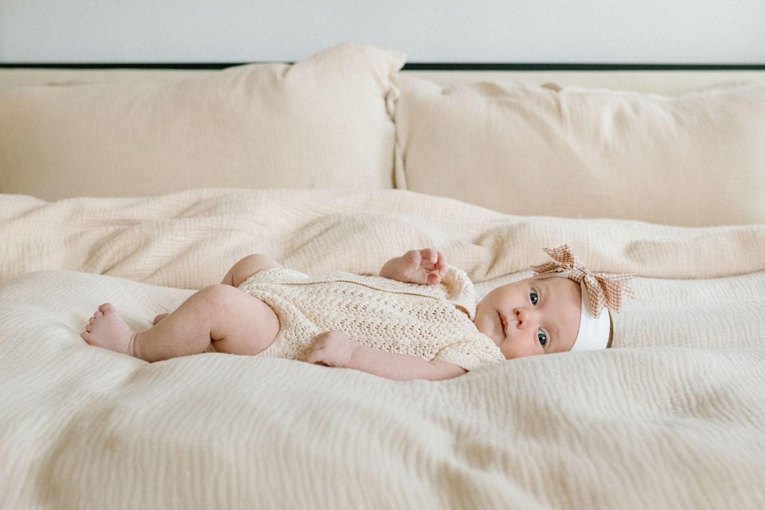 Adorable baby in cream knit outfit and bow headband lying on a soft beige bed.