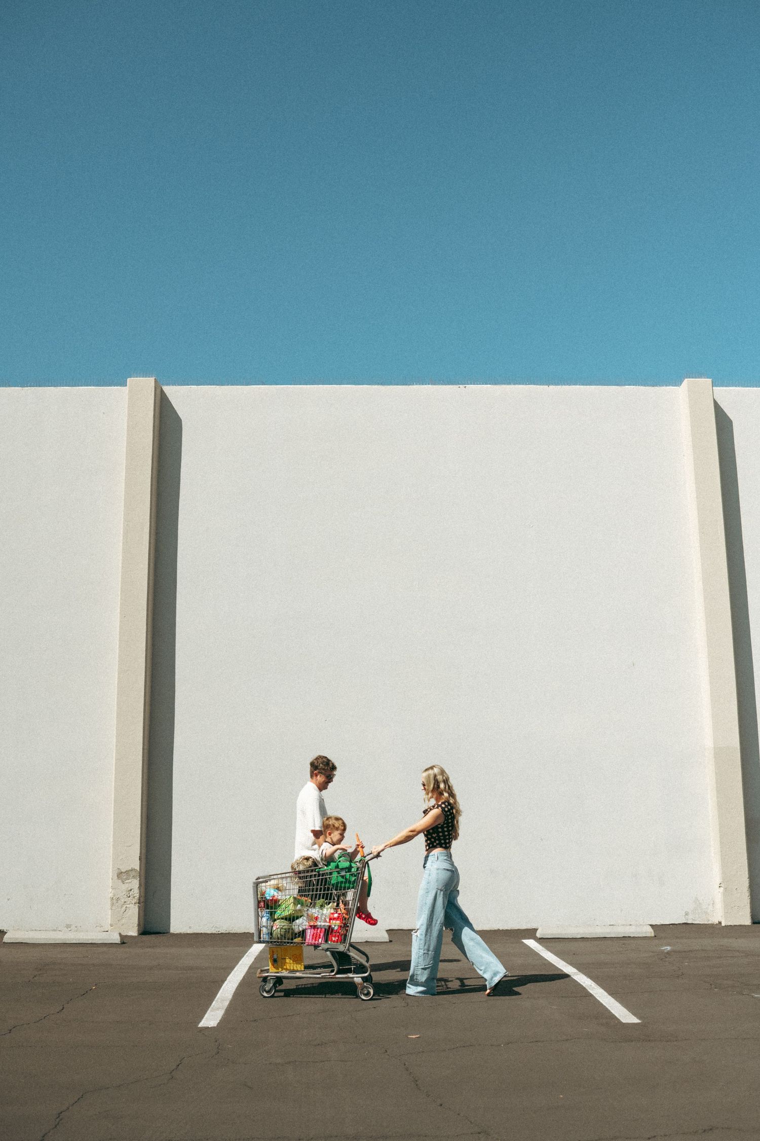 Family with child in shopping cart walks through sunny parking lot against white wall and blue sky.