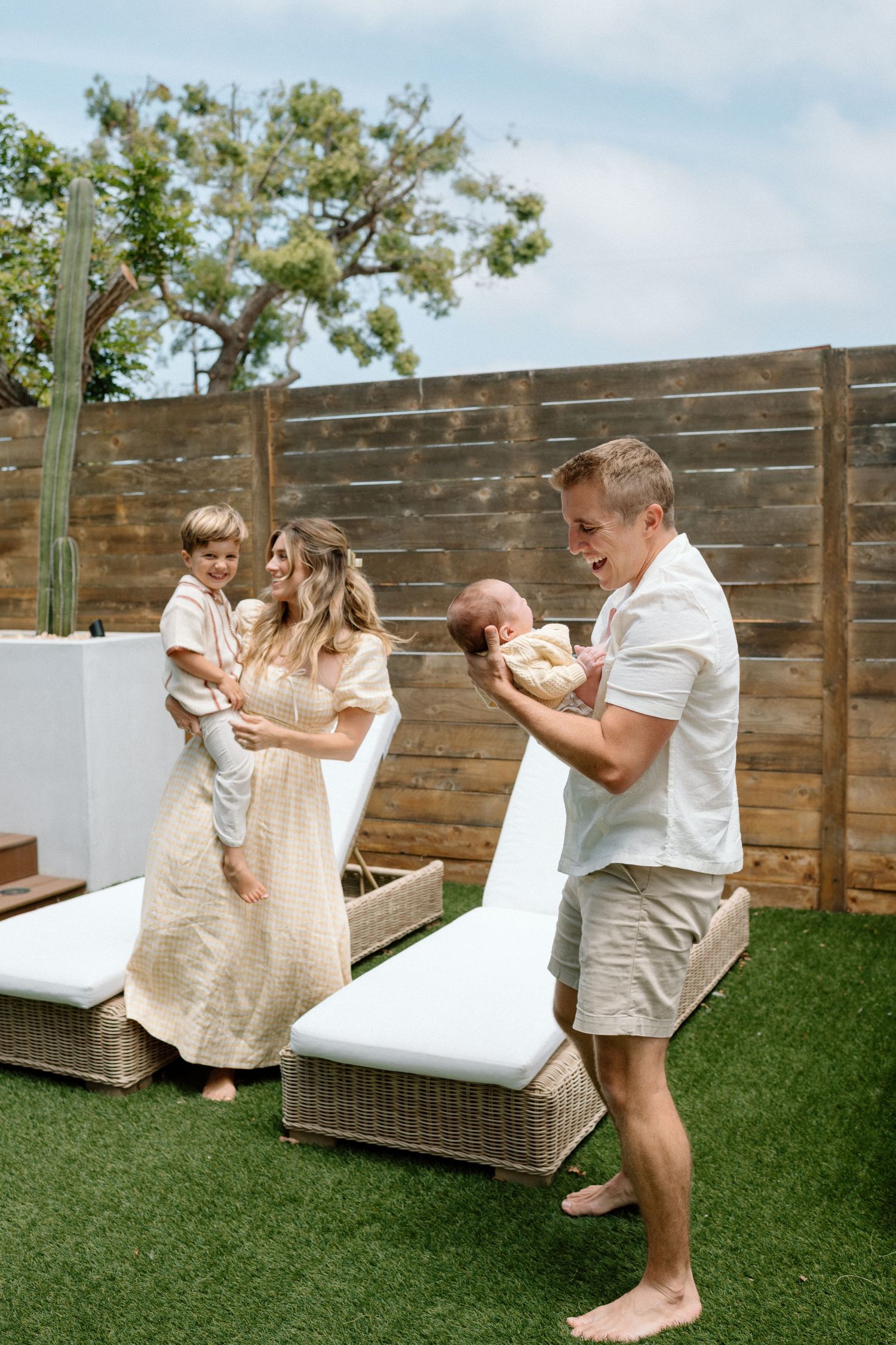 A happy family plays in a sunny backyard, with parents holding a toddler and newborn baby.