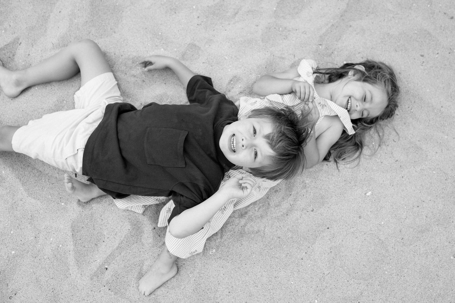 Two smiling children lie together on sandy beach in a black and white photo.
