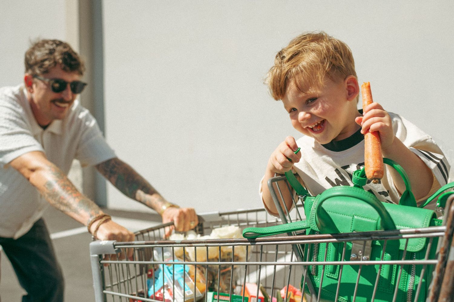 Young boy sitting in a shopping cart holding a carrot while a tattooed man pushes the cart outside a store.