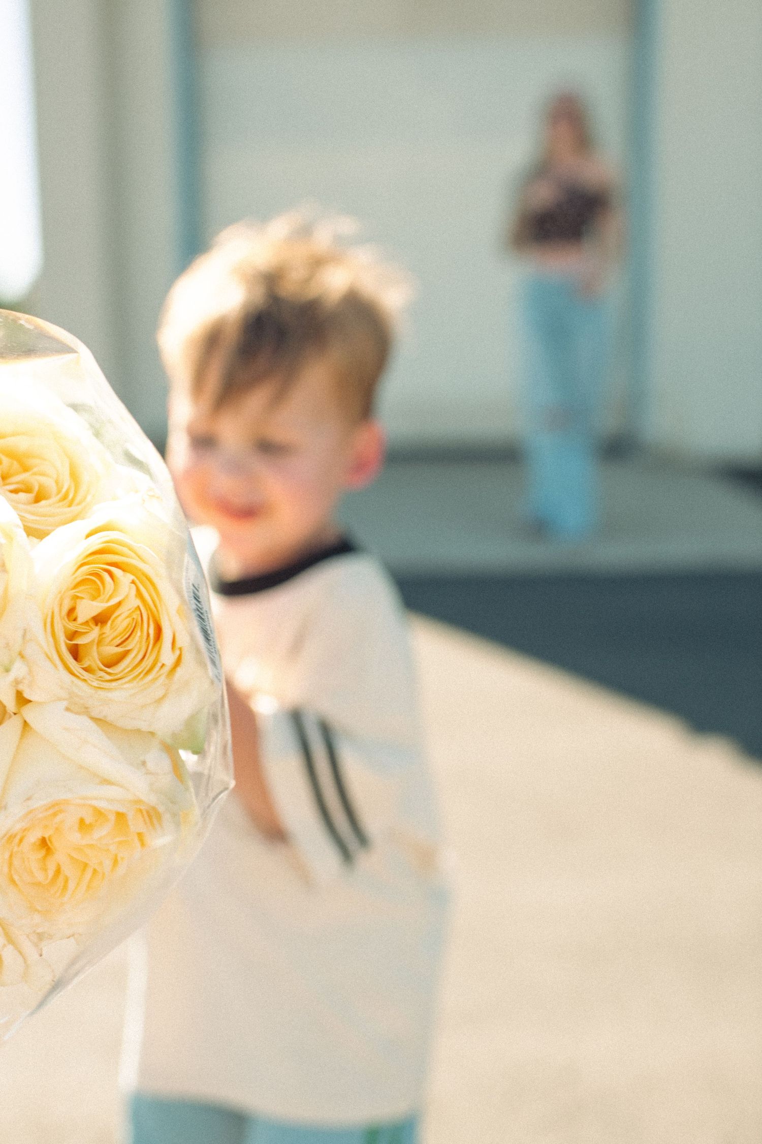 A young boy holds a bouquet of yellow roses toward the camera, with a woman standing in the background.