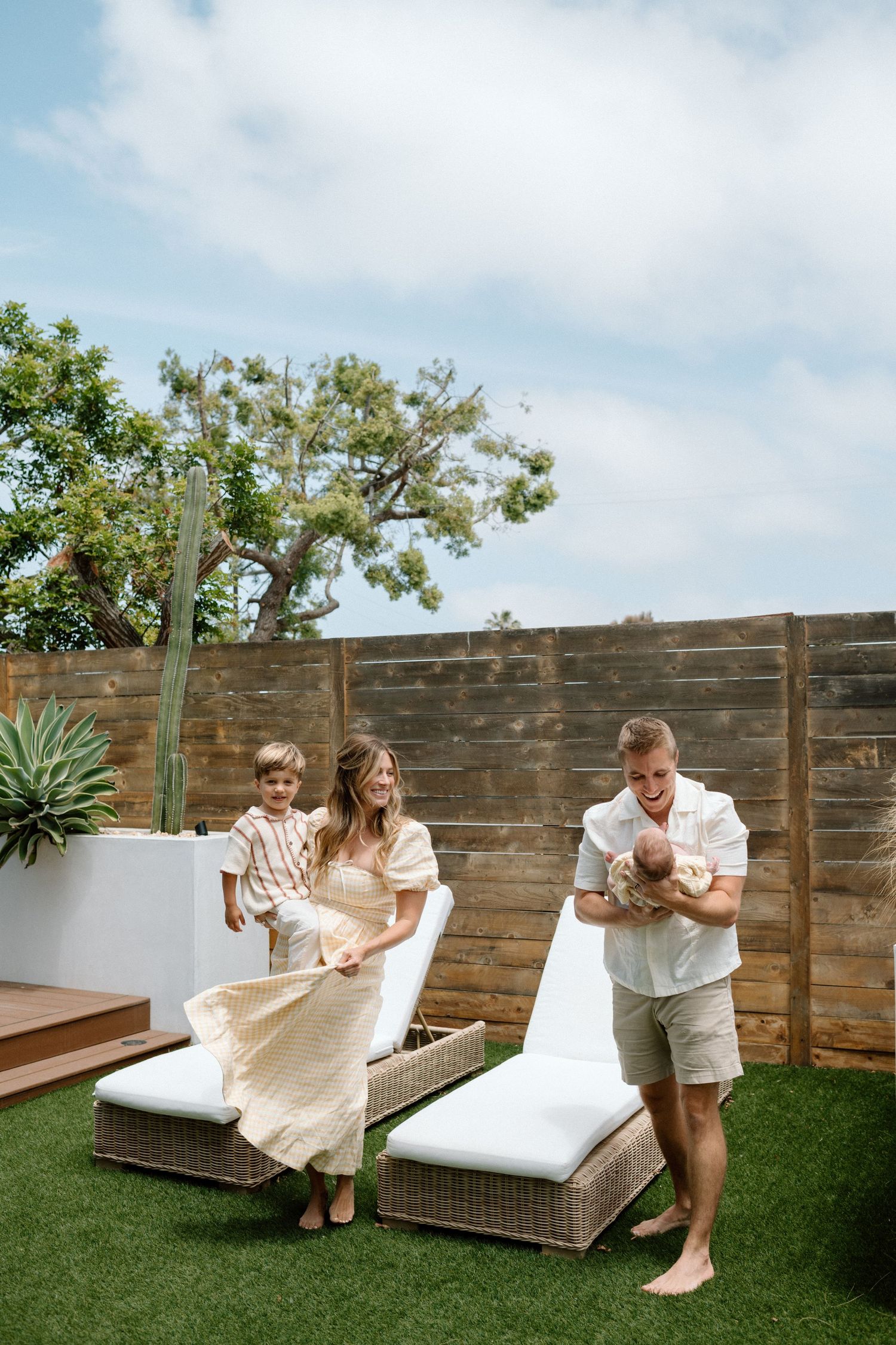 Young family with toddler and newborn relaxing in a sunny backyard with lounge chairs.