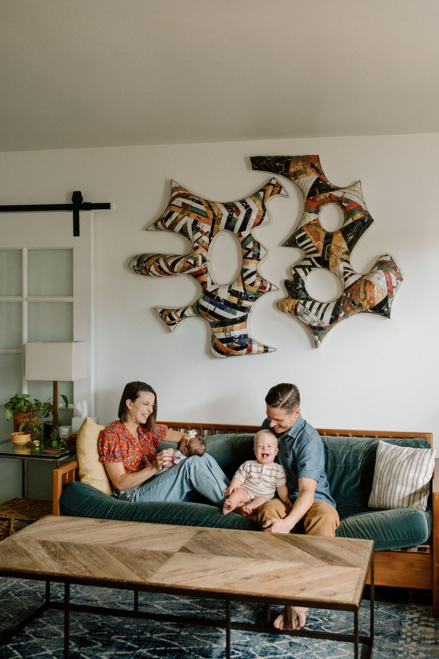 Young family with two babies enjoying quality time together on a teal sofa beneath colorful wall art.