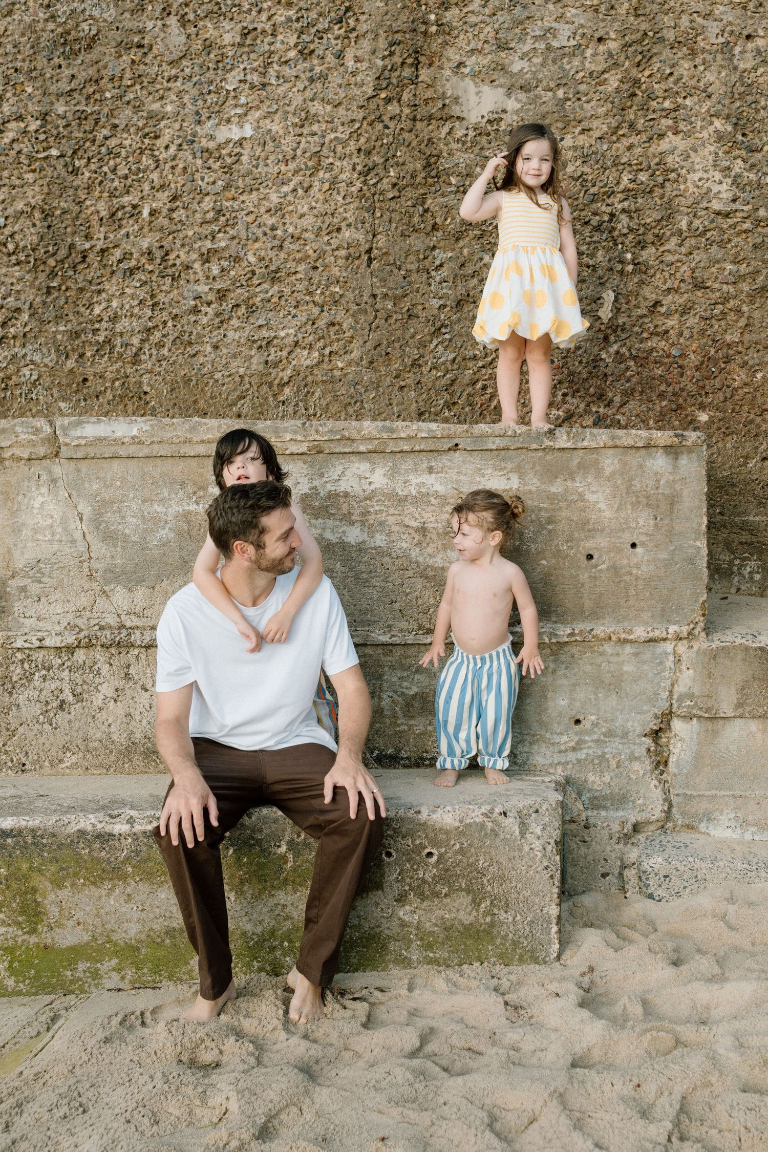 Father in white tee sits on sandy stone steps with three young children climbing around him.