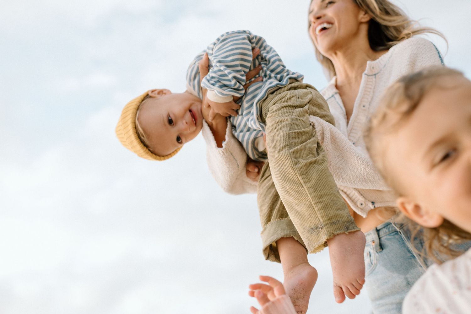 A joyful mother lifts her toddler in a striped shirt outdoors while another child plays nearby under a bright sky.
