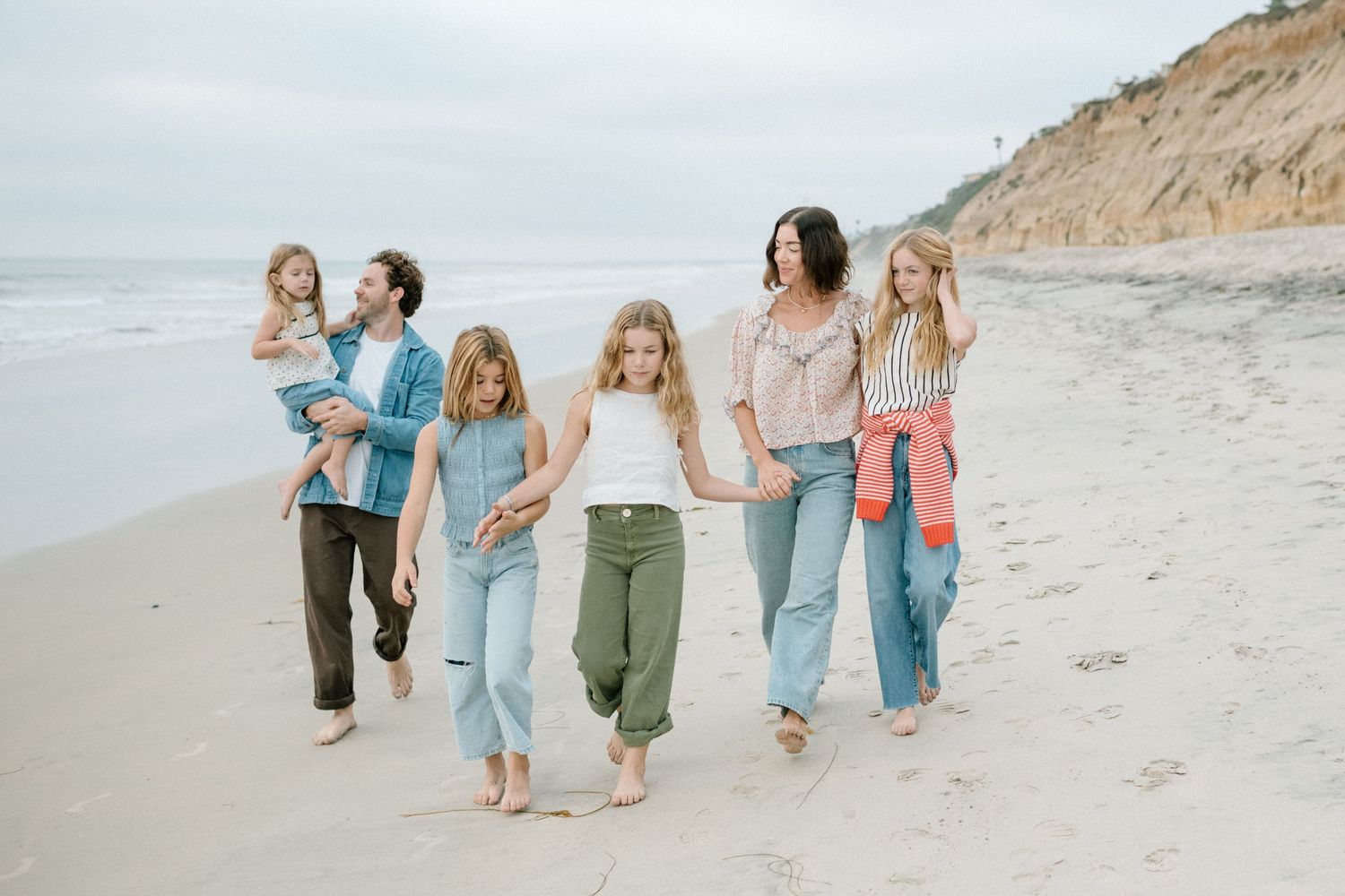 A family of six walking hand in hand along a sandy beach with towering cliffs and overcast skies behind them.