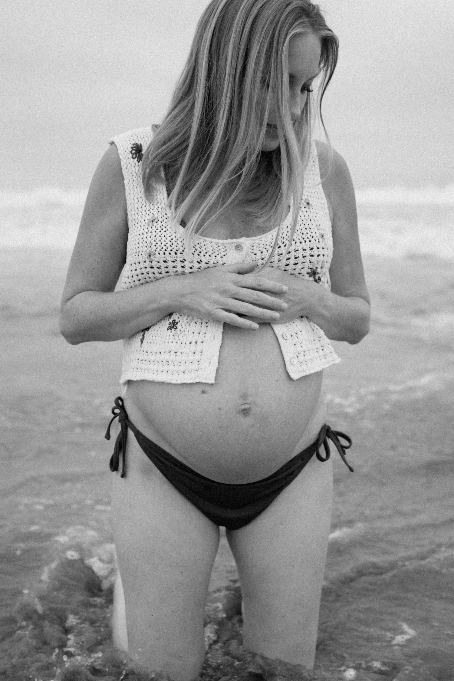 Black and white photo of pregnant woman in bikini and crochet top cradling baby bump at beach.