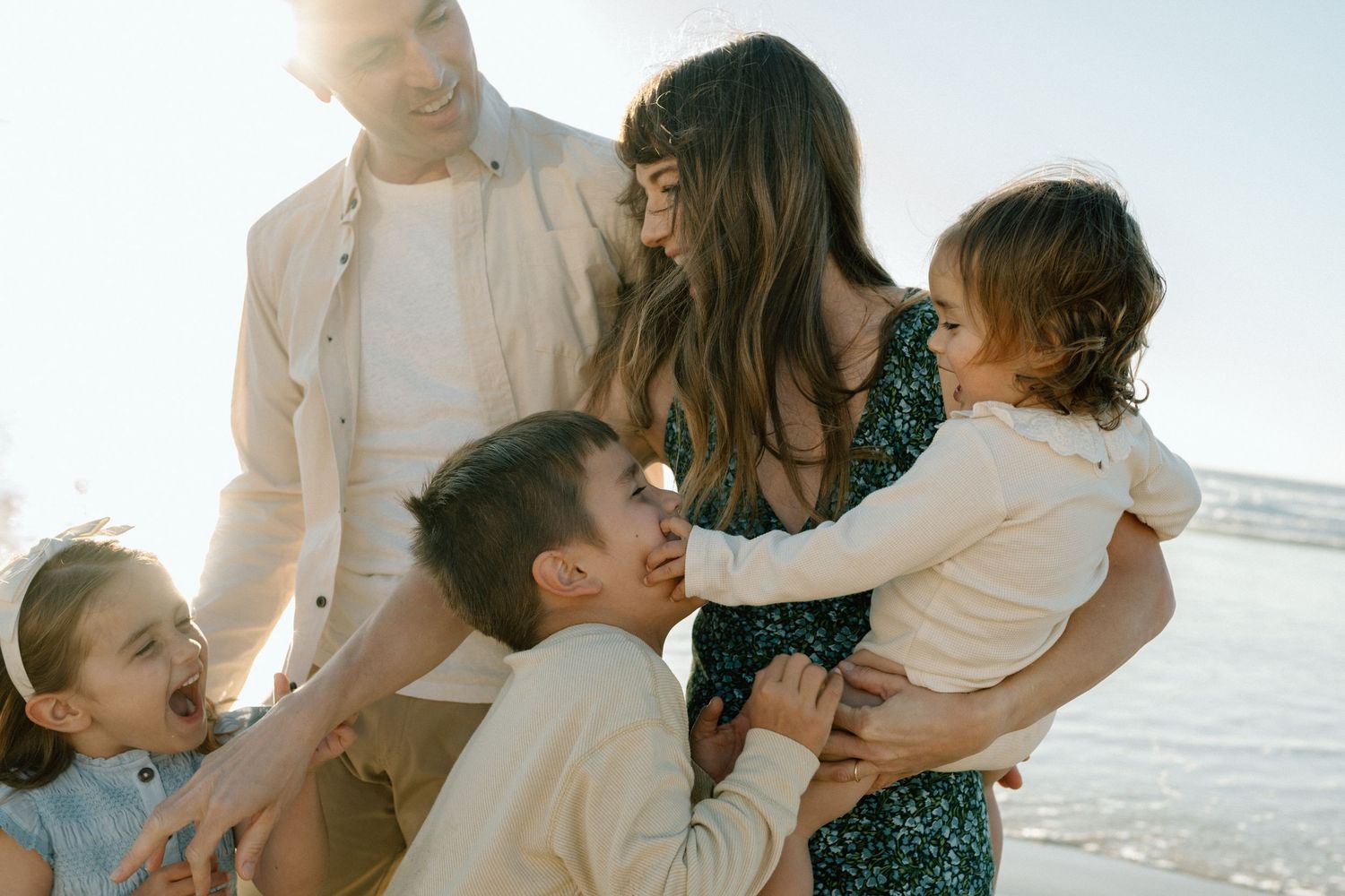 A happy family of five shares a warm embrace on a sunny beach with shimmering water behind them.