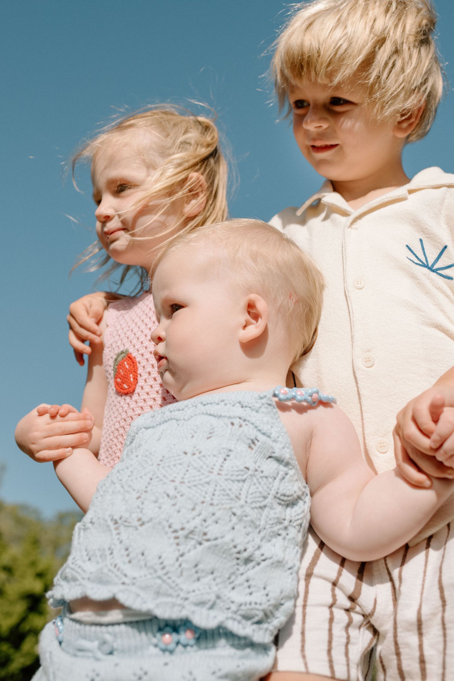 Three young blonde siblings standing together outdoors under a bright blue sky.