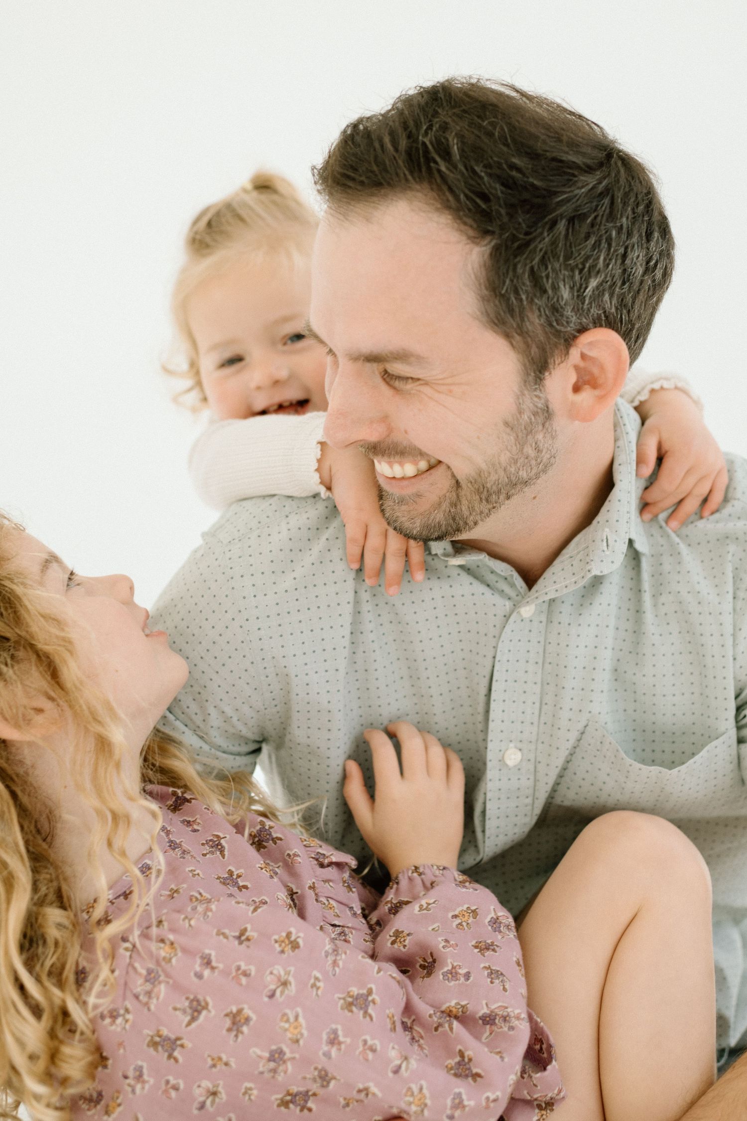 Bearded father laughs joyfully while holding toddler on shoulders as older daughter looks up smiling.