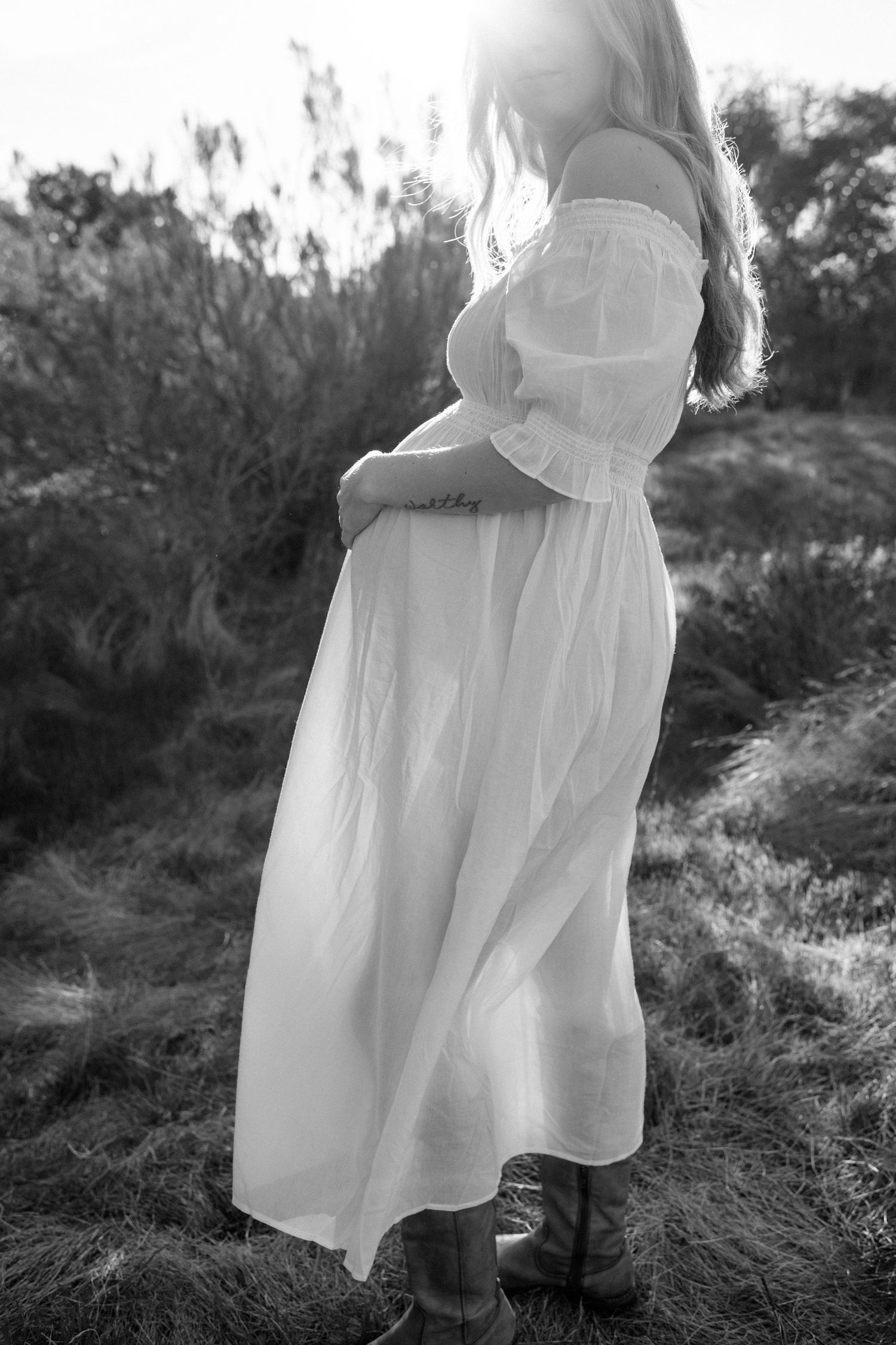 Black and white silhouette of pregnant woman in flowing white off-shoulder dress outdoors.