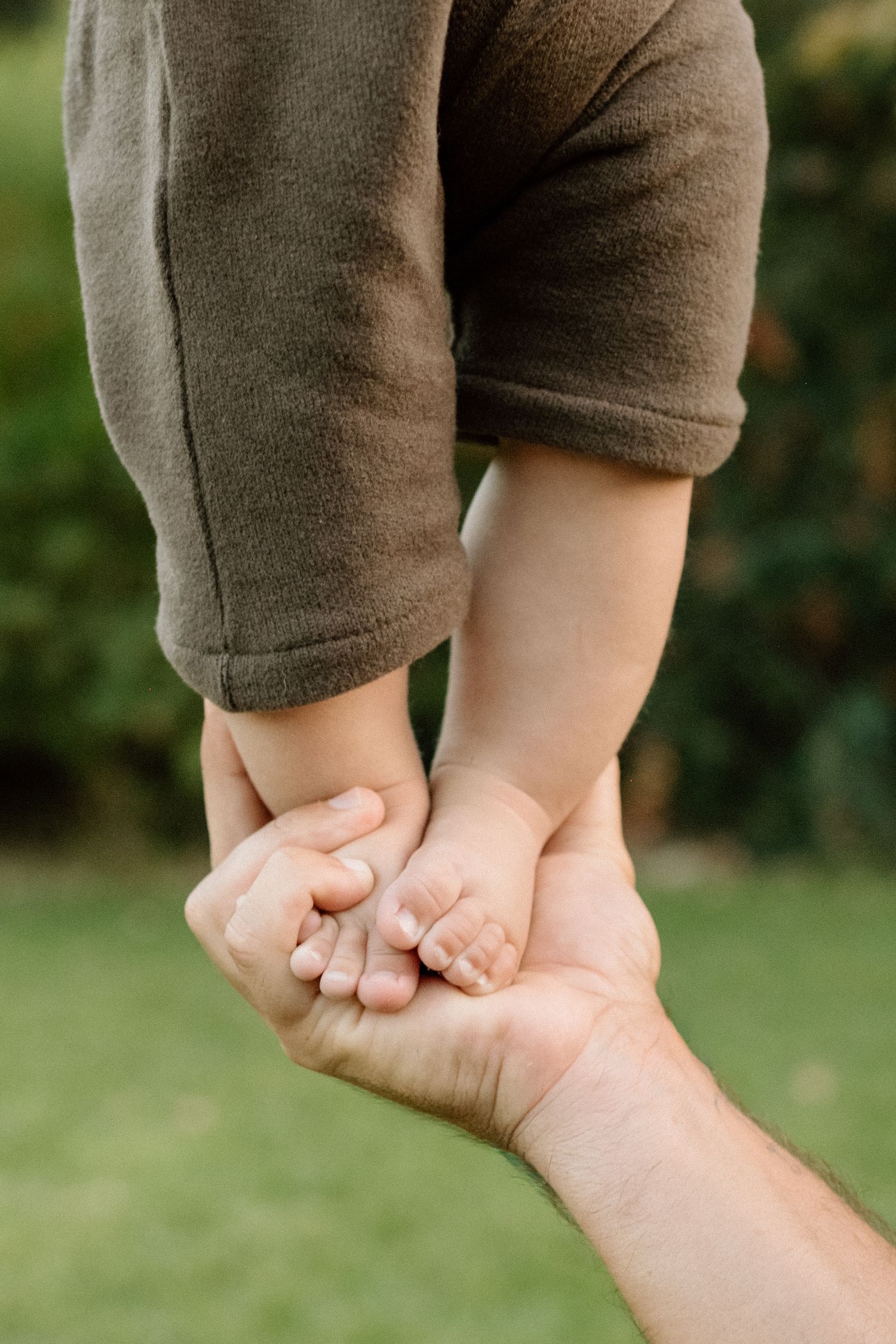 Adult hands gently holding tiny baby feet outdoors on green grass background.
