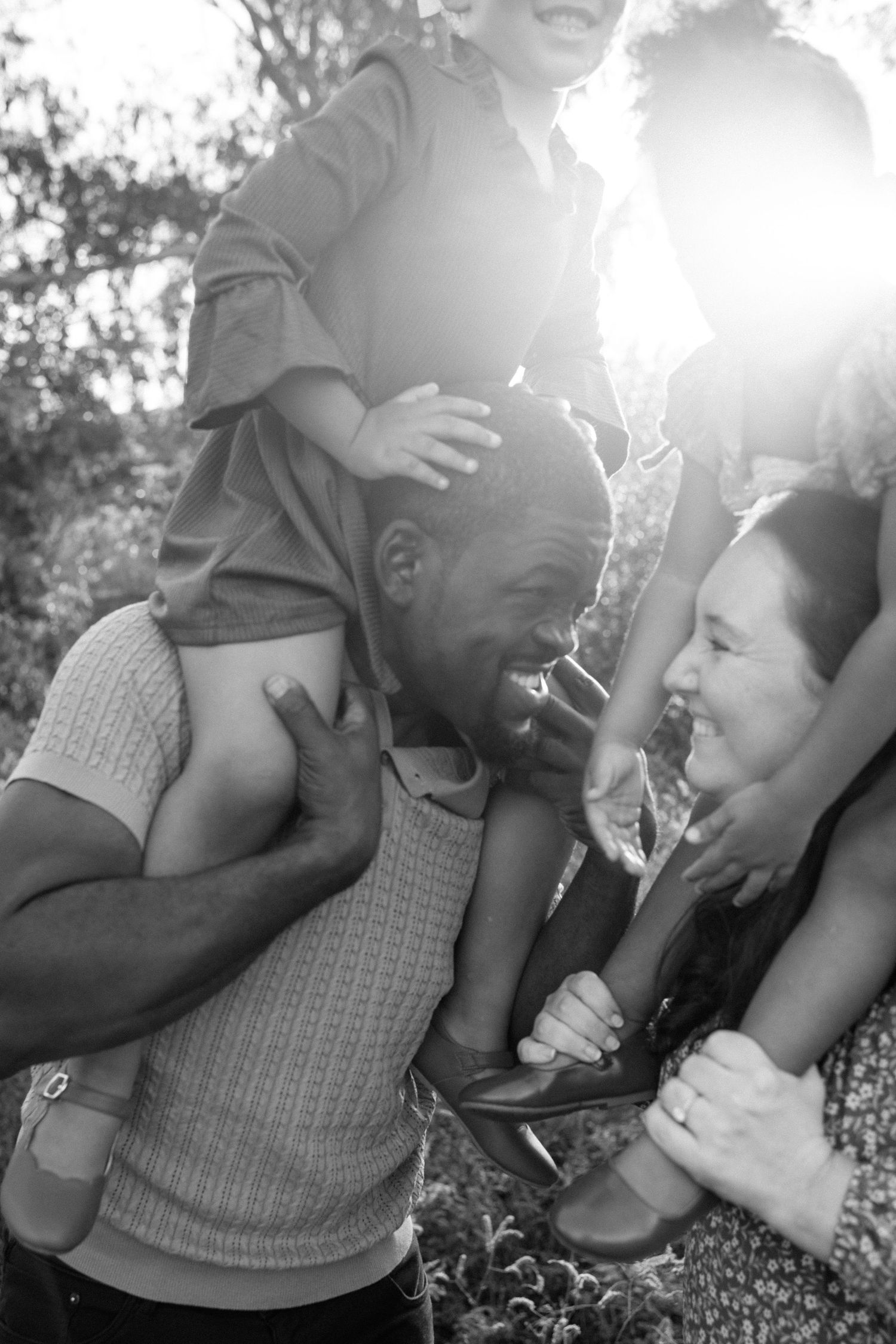 Black and white photo of laughing multiracial family with child on fathers shoulders in sunny backyard.
