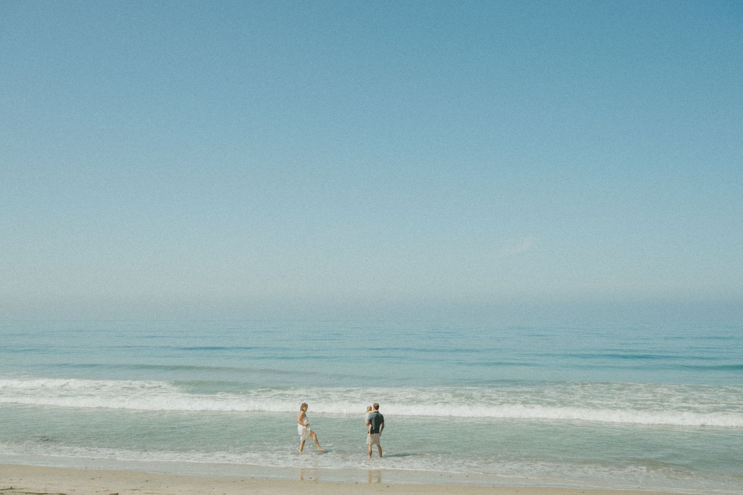 Two children playing at the ocean shoreline on a sunny day with calm blue waves and clear sky.