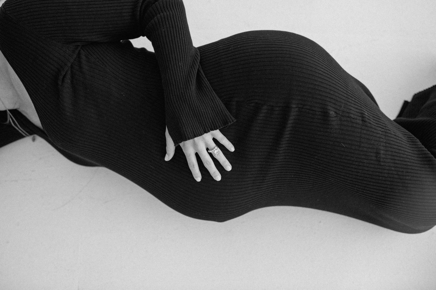 Black and white artistic photo of pregnant woman in black ribbed dress lying on white surface.