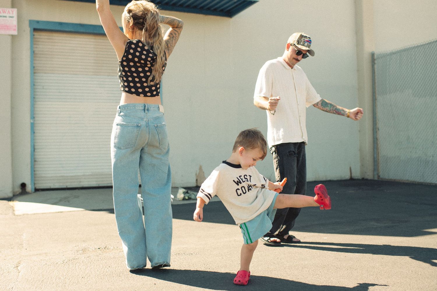 Family dancing outdoors, woman in crop top and wide-leg jeans, child in white tee, man in cap and jeans.