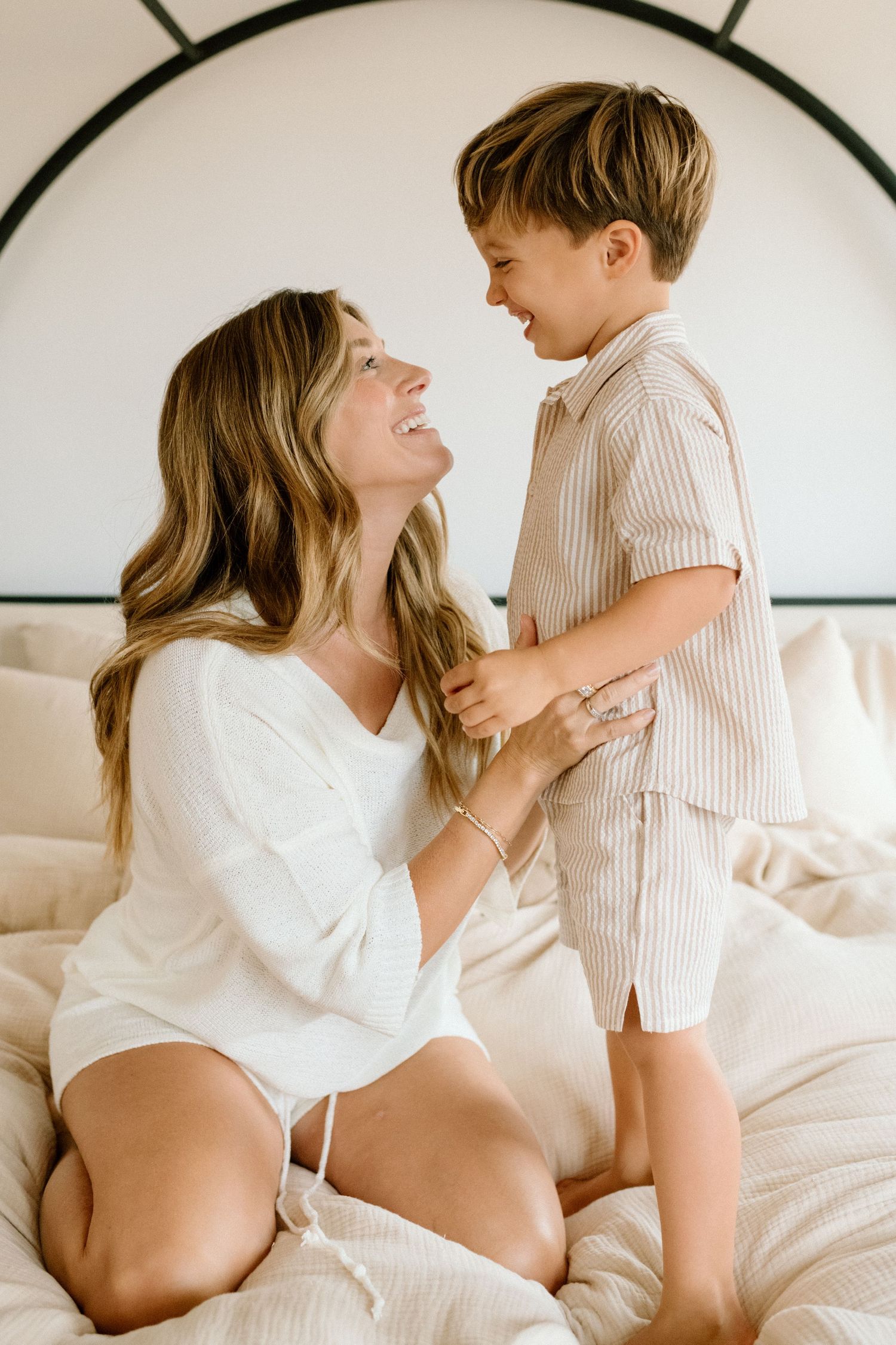 Joyful mother and young son smiling at each other while playing on a white bed.