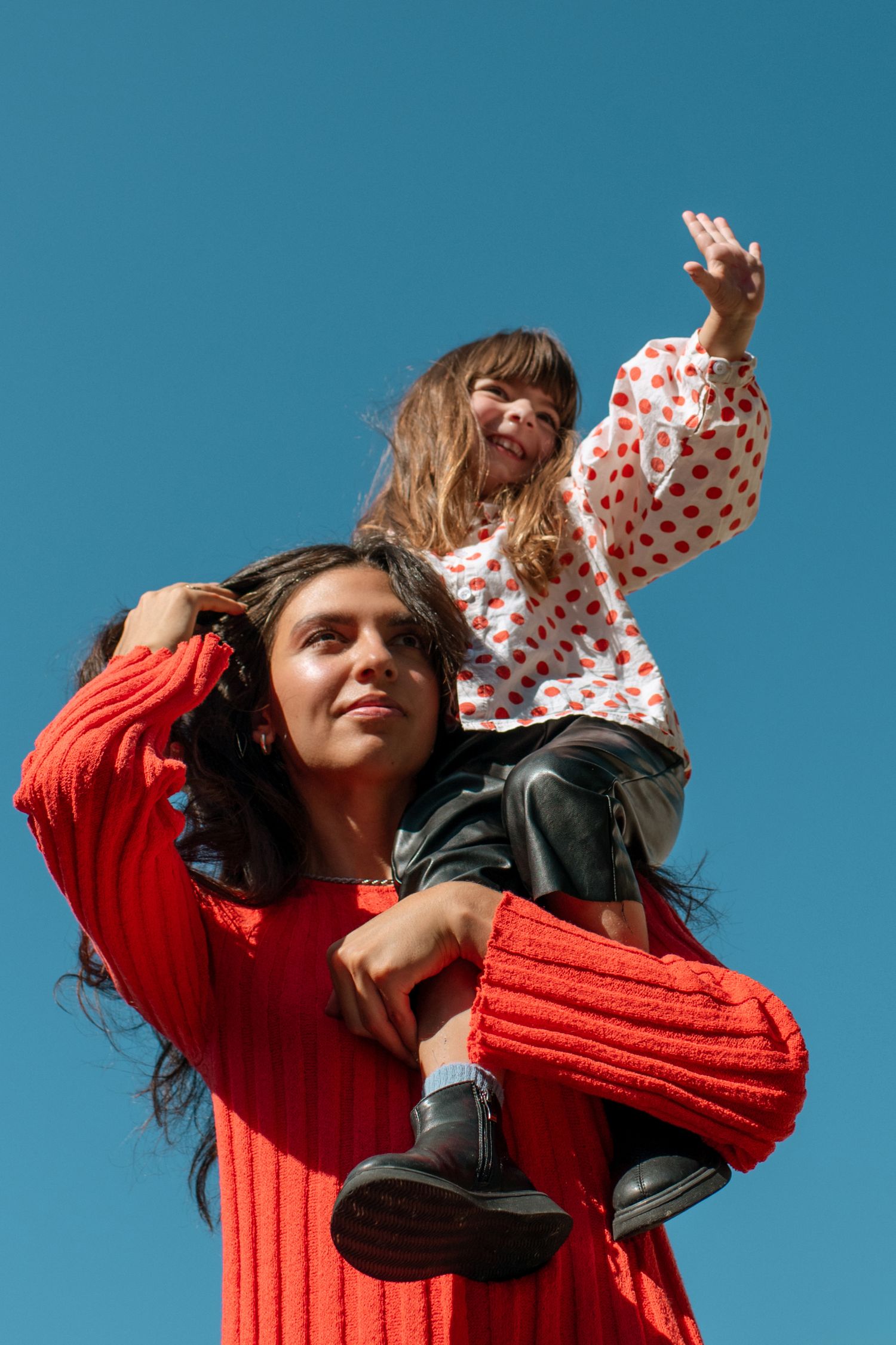 Woman in red sweater carrying a young girl in polka dot shirt on her shoulders against a blue sky.