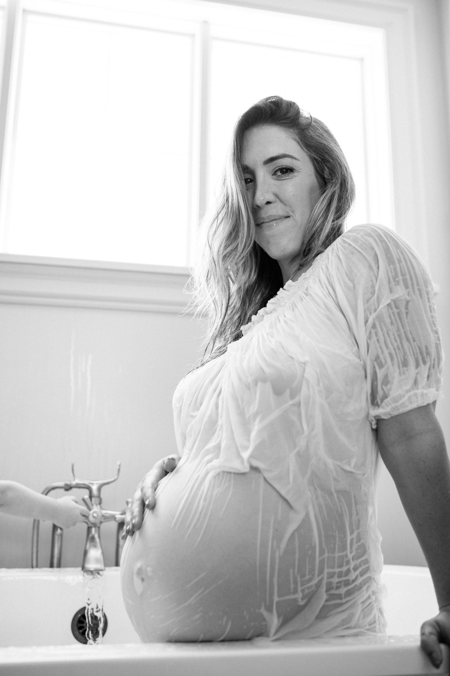 Black and white photo of a pregnant woman in a wet white dress sitting in a bathtub, smiling at the camera.