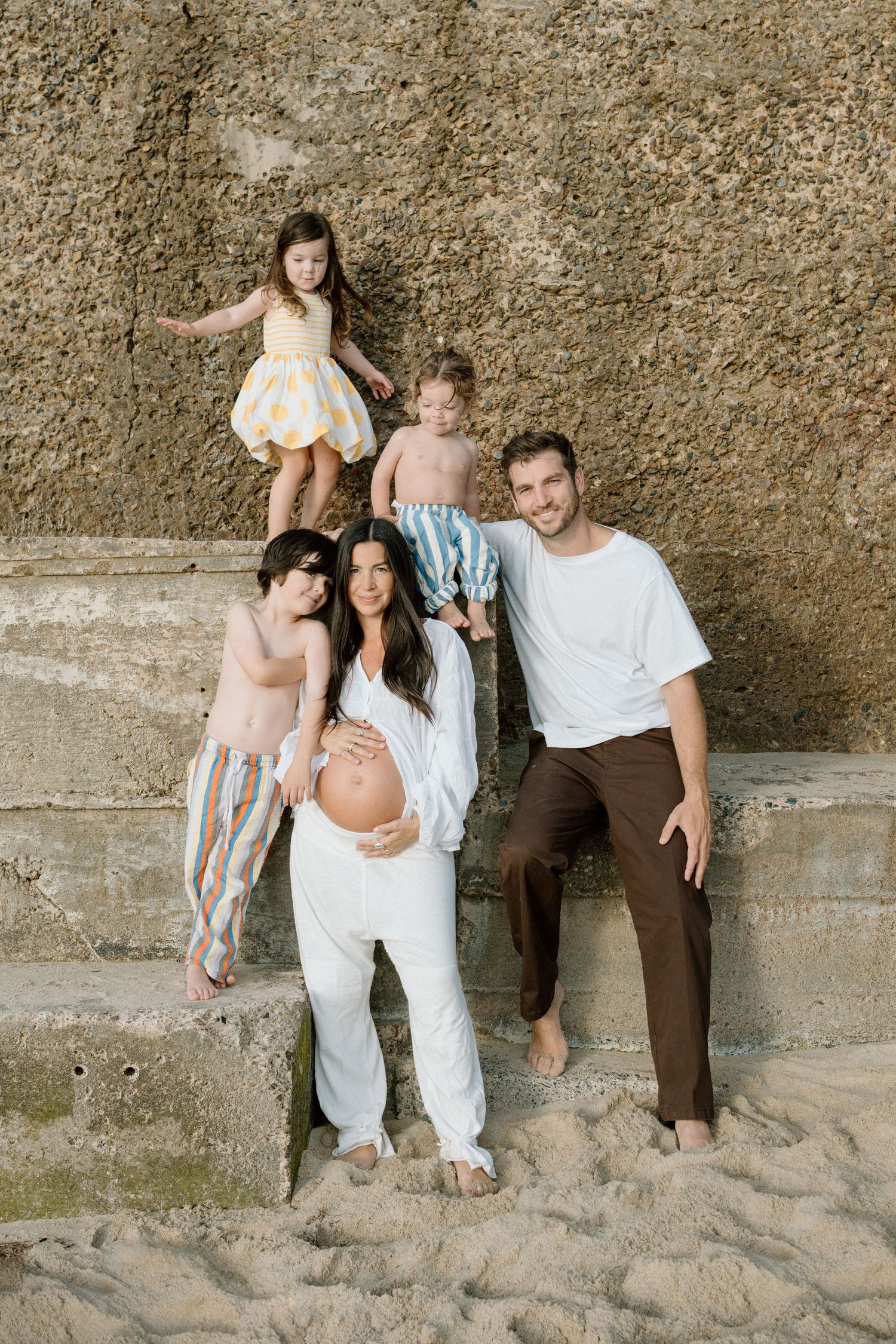 A smiling pregnant family of five poses together on stone beach steps in warm natural light.