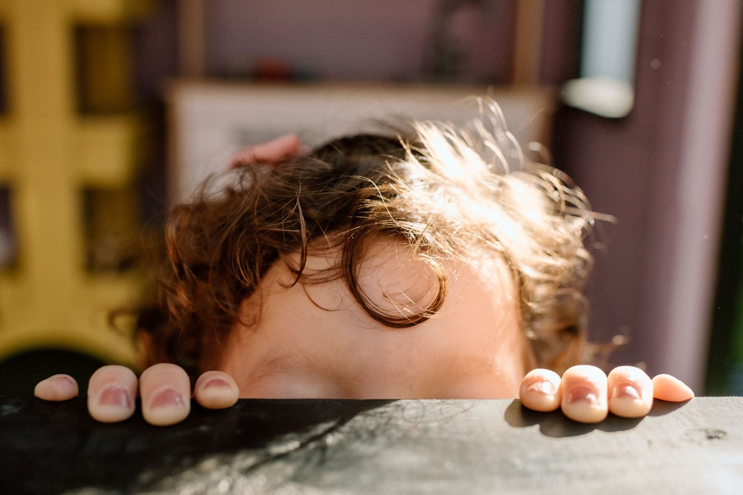 Curious curly-haired child peeking over the edge of a table with tiny fingers.