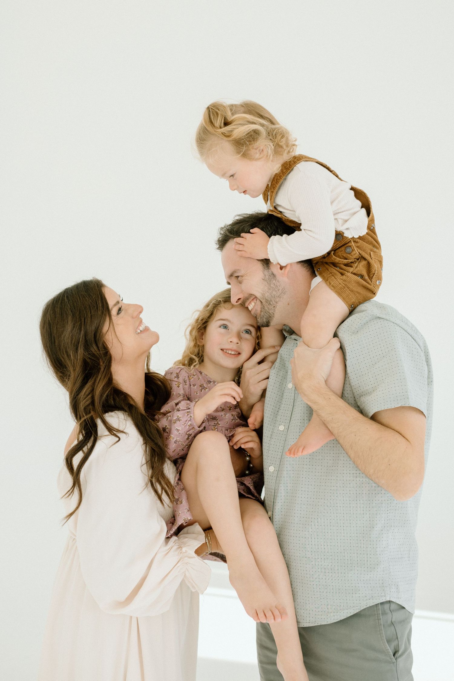 Happy family of four with father carrying toddler on shoulders while mother and daughter smile together.