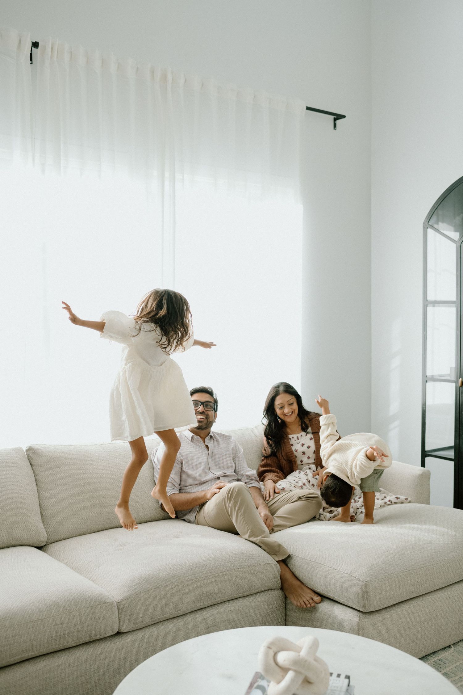 Family enjoying playful time on a white sofa as girl jumps in bright room.