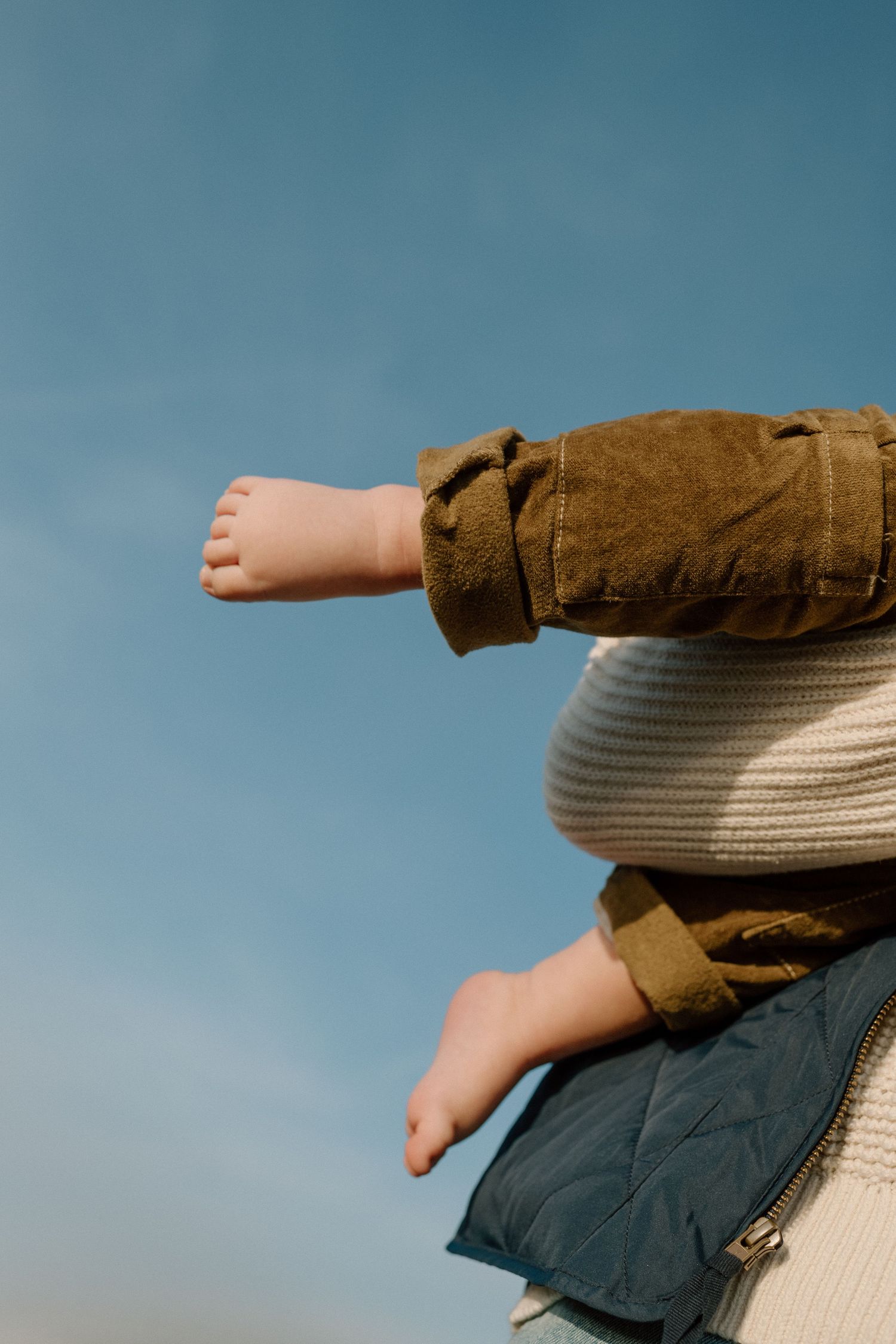 Close-up of a babys bare feet and legs in olive pants, held against an adults chest under a blue sky.