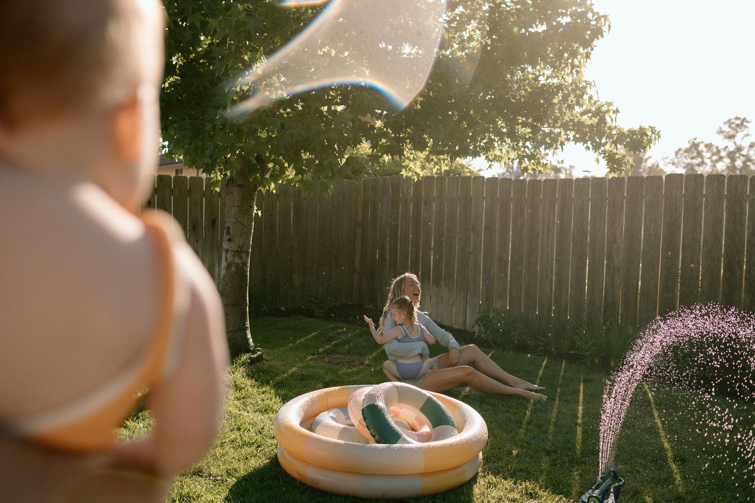 Child playing with a sprinkler near an inflatable pool in a sunny backyard.