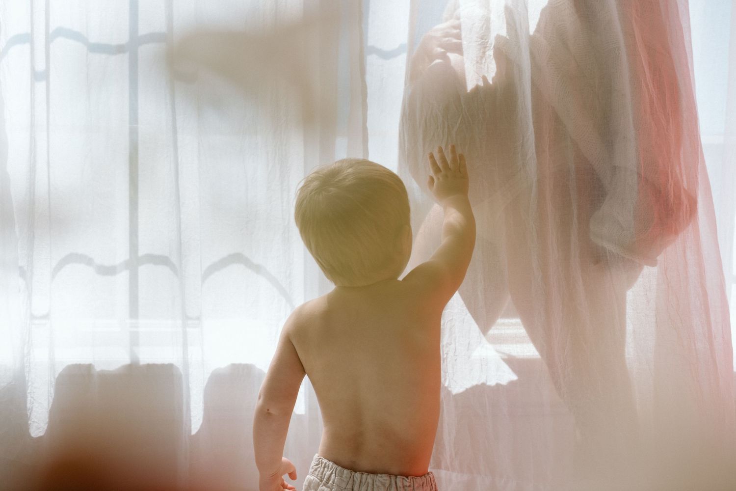 A shirtless toddler reaches up toward a pregnant woman standing by sheer white curtains in soft natural light.